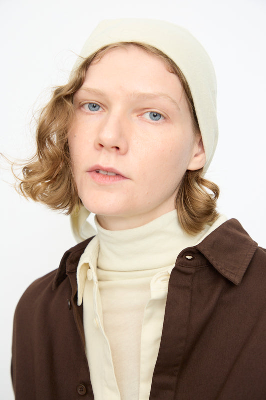 A person with short wavy hair wears the Black Crane Triangle Scarf in Natural, crafted from a soft cotton cashmere blend, styled with a cream turtleneck and brown shirt, gazing at the camera against a plain white background.