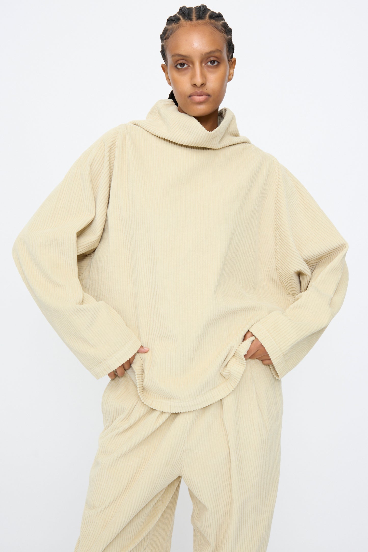 A person wears the Black Crane Tube Top in Ivory, a loose, boxy-fit cotton corduroy top with long sleeves and matching pants, standing against a plain white background with hands in pockets.