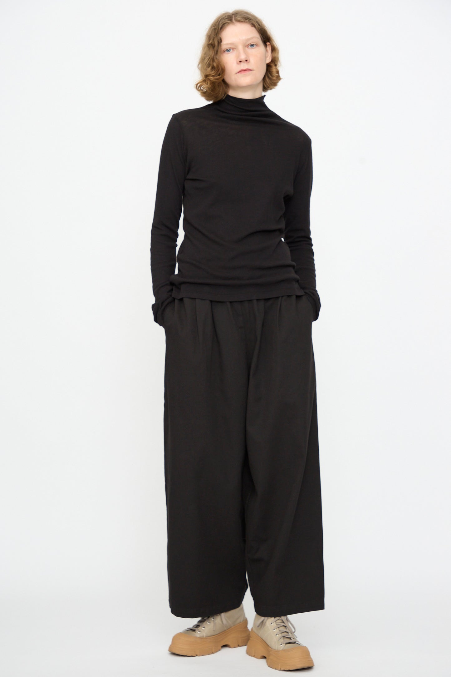 A person stands against a white background, dressed in the Black Crane Turtleneck in Black and wide black relaxed-fit pants, paired with beige platform shoes. Their hands are in their pockets and they have a neutral expression.