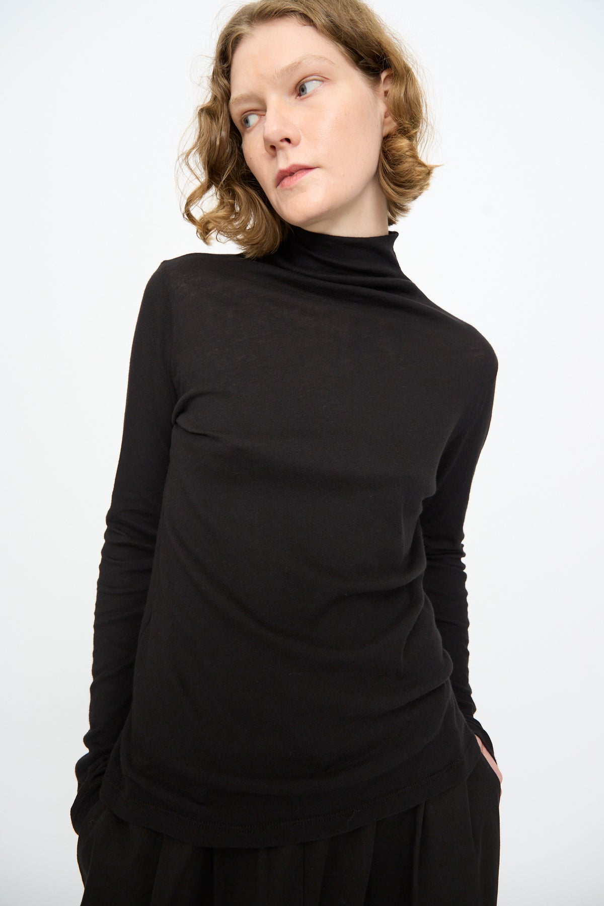 A person with short, wavy blond hair wears the Black Crane Turtleneck in Black—a relaxed fit cotton-cashmere top—standing against a plain white background and looking to the side with hands in pockets.