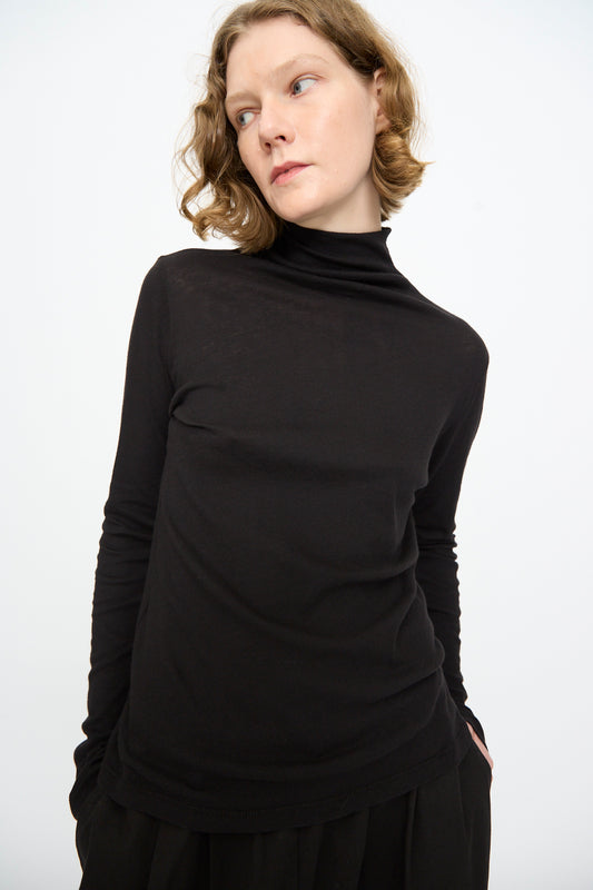 A person with short, wavy blond hair wears the Black Crane Turtleneck in Black—a relaxed fit cotton-cashmere top—standing against a plain white background and looking to the side with hands in pockets.