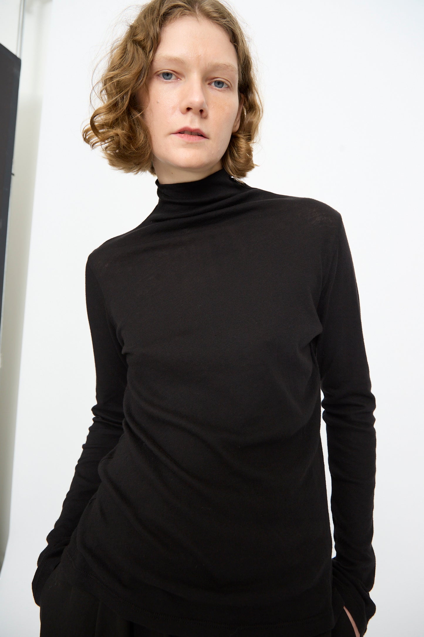 A person with short curly hair stands against a plain white background, looking directly at the camera with a neutral expression, wearing the Black Crane Turtleneck in Black in a relaxed fit.