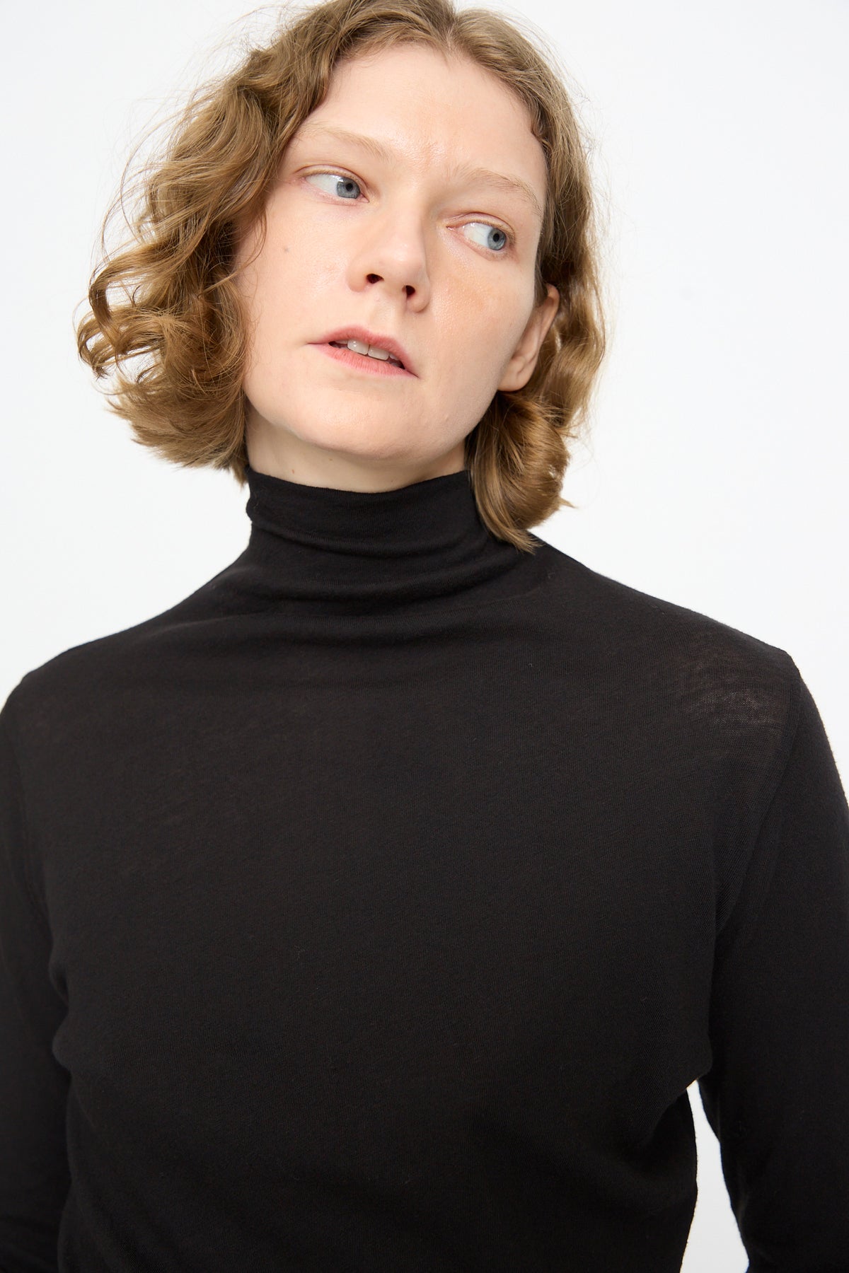A person with short, wavy light brown hair wears the Black Crane Turtleneck in Black, featuring a relaxed fit in cotton cashmere, and gazes slightly to the side against a plain white background.