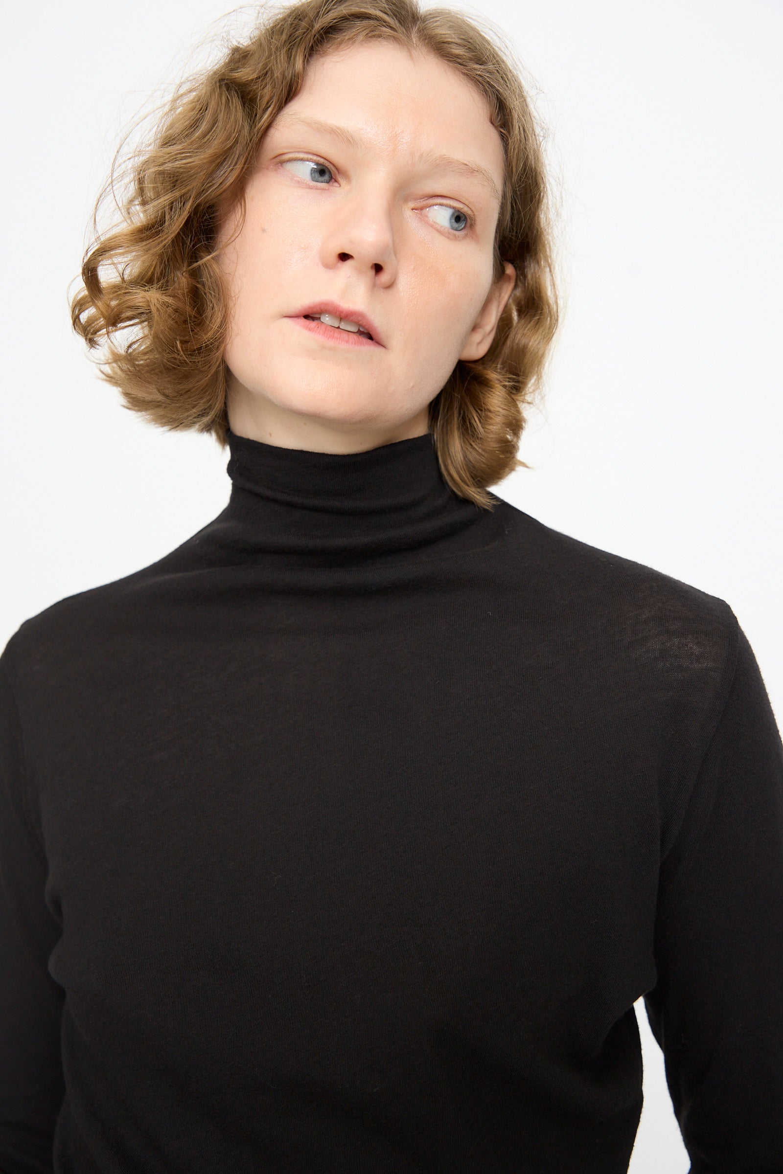 A person with short, wavy light brown hair wears the Black Crane Turtleneck in Black, featuring a relaxed fit in cotton cashmere, and gazes slightly to the side against a plain white background.
