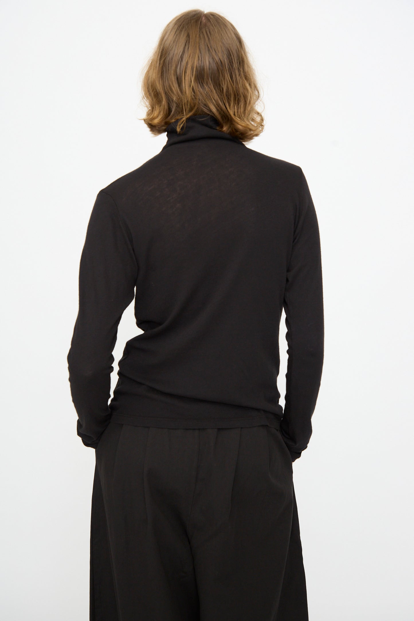 A person with shoulder-length hair is shown from the back against a plain white background, wearing the Black Crane Turtleneck in Black—a relaxed fit cotton-cashmere top—paired with black pants.