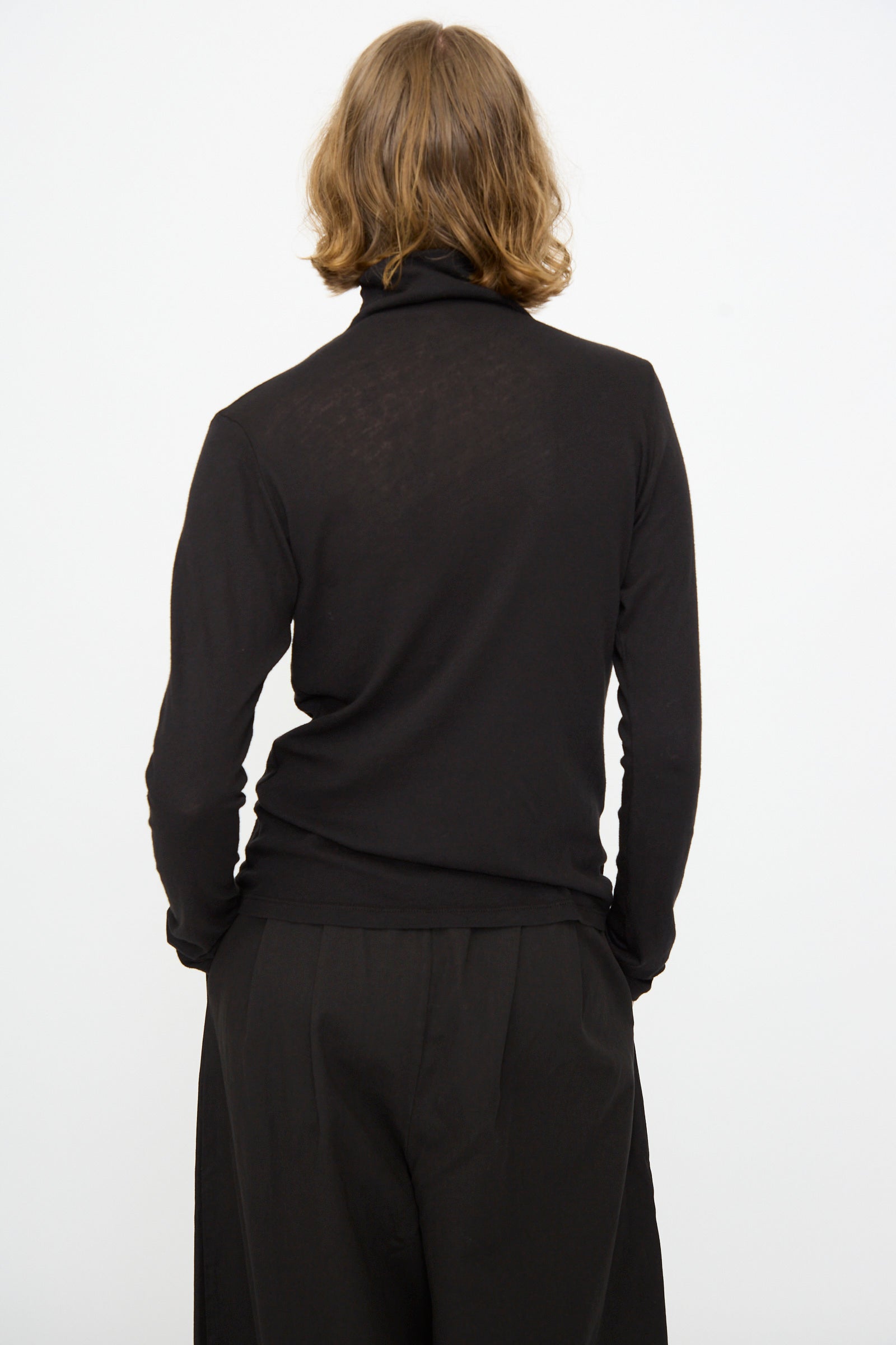 A person with shoulder-length hair is shown from the back against a plain white background, wearing the Black Crane Turtleneck in Black—a relaxed fit cotton-cashmere top—paired with black pants.