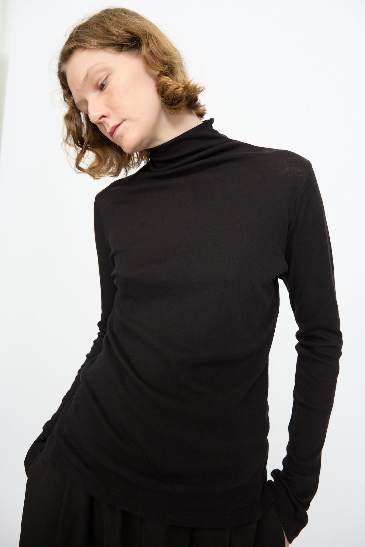 A person with short, wavy hair wears the Black Crane Turtleneck in Black—a relaxed-fit, long-sleeve cotton cashmere top—standing against a plain white background with hands in pockets, looking down.