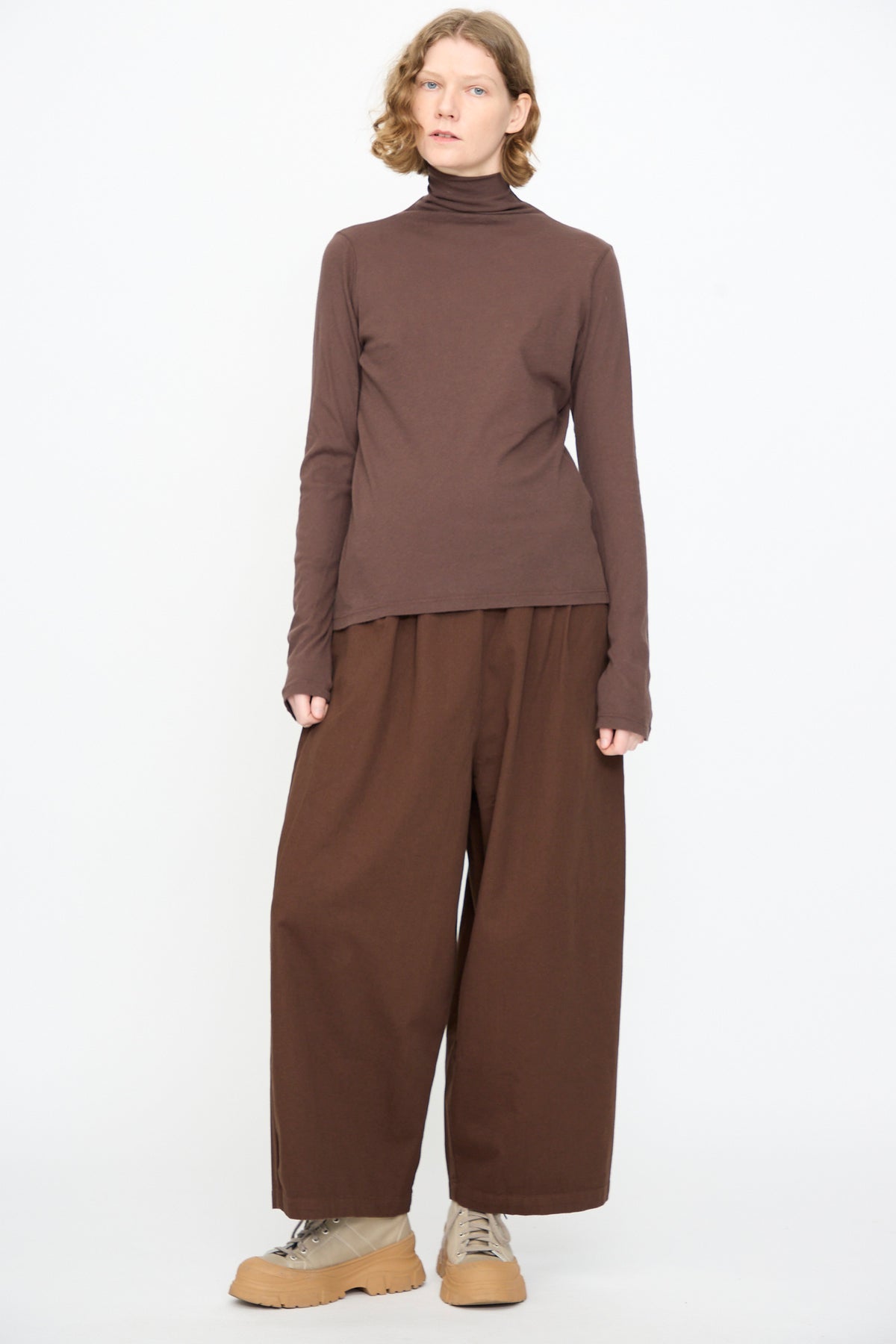 A person stands against a white background wearing the Black Crane Turtleneck in Dark Plum, featuring a relaxed fit in a soft cotton cashmere blend, styled with wide brown pants and beige shoes, with a neutral facial expression.