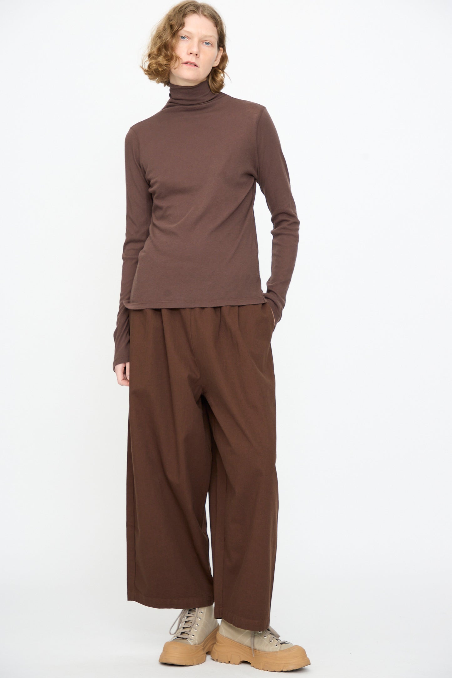 A person wearing the Black Crane Turtleneck in Dark Plum, paired with loose brown pants and beige shoes, stands against a plain white background with a neutral expression.