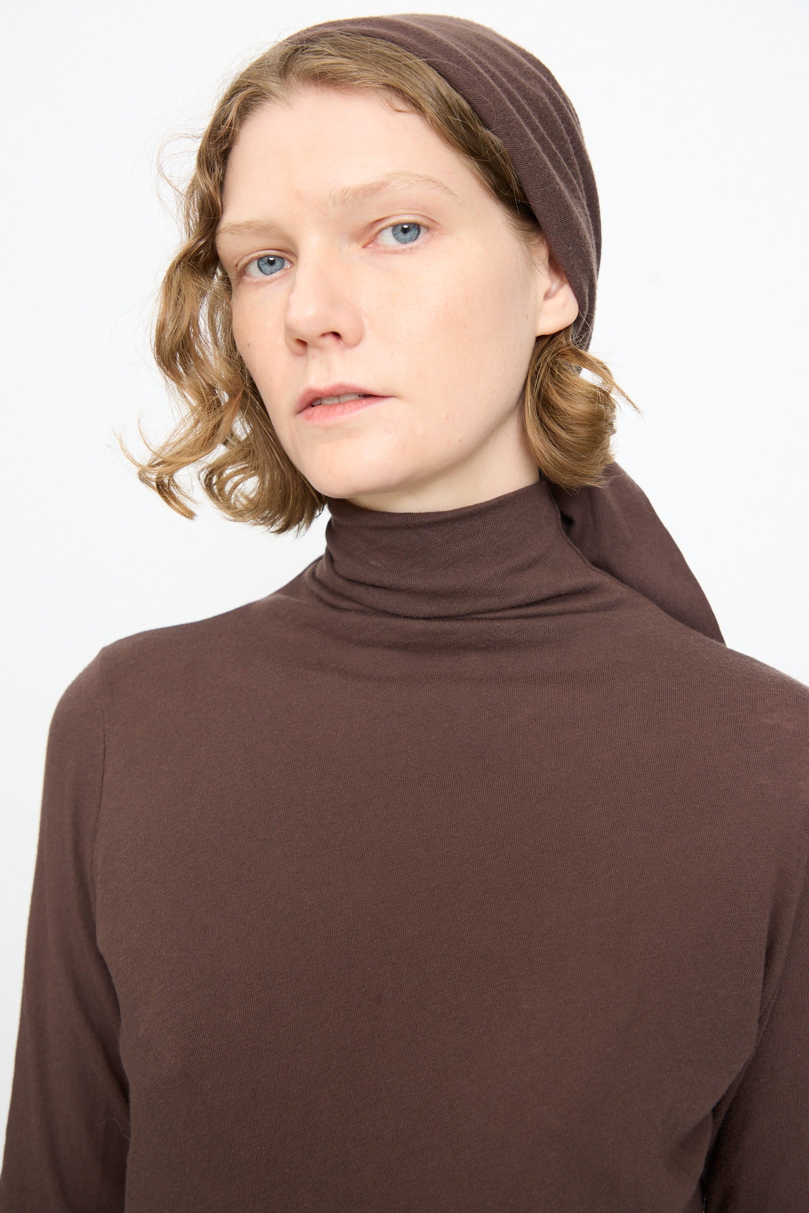 Person with short wavy hair wearing the Black Crane Turtleneck in Dark Plum and a matching headscarf, looking at the camera with a neutral expression against a plain background.