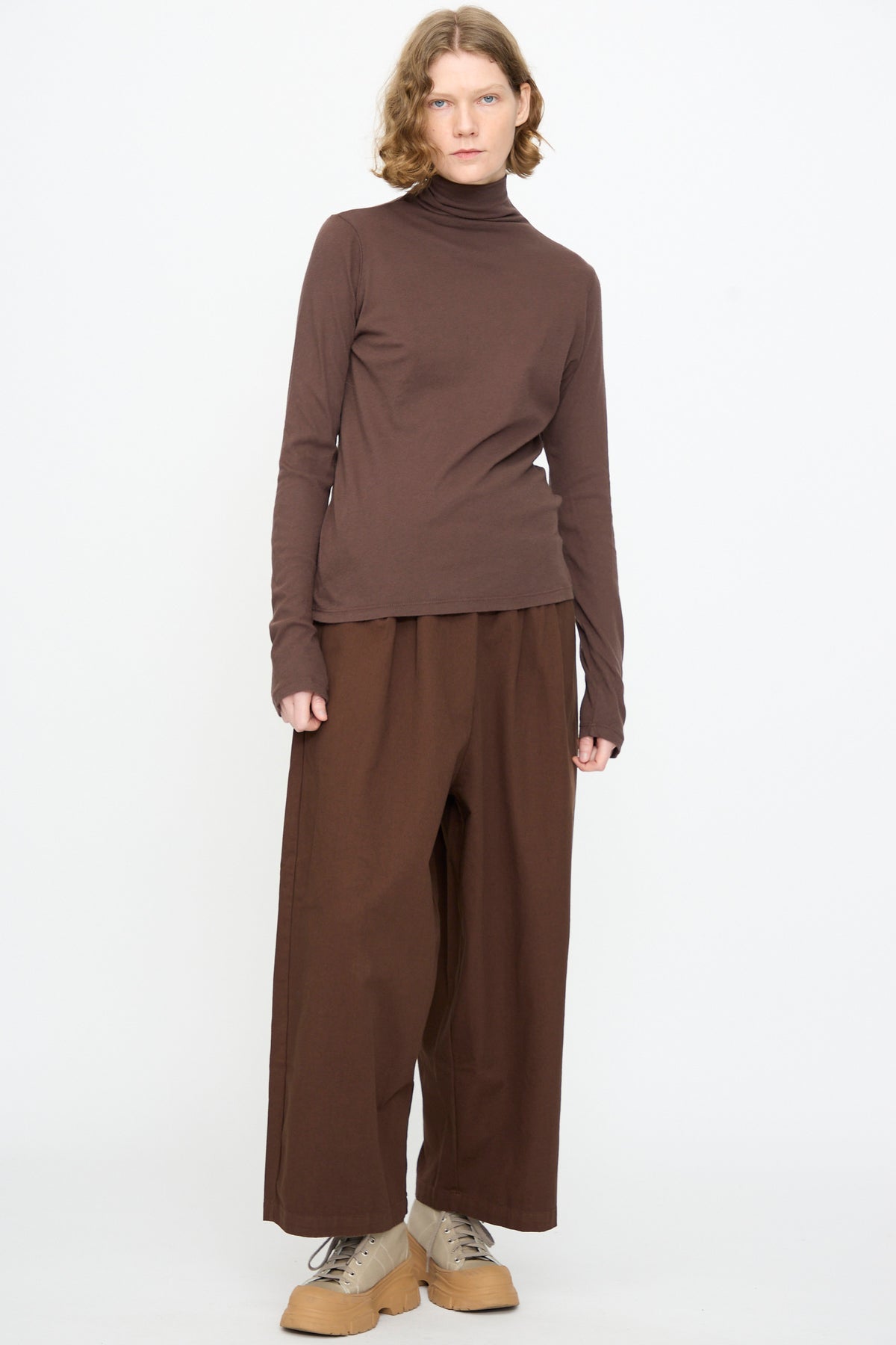 A person wearing the Black Crane Turtleneck in Dark Plum, featuring a relaxed fit and soft cotton cashmere blend, paired with brown wide-leg pants and tan platform shoes, stands against a plain white background.
