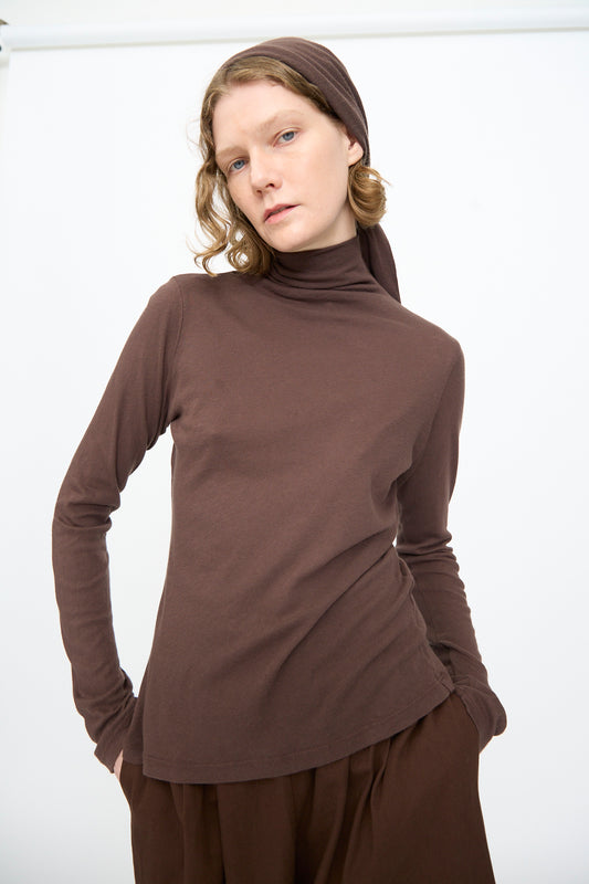 A person wearing the Black Crane Turtleneck in Dark Plum, styled in a relaxed fit with a matching headscarf, stands against a plain white background, looking directly at the camera with hands in their pockets.