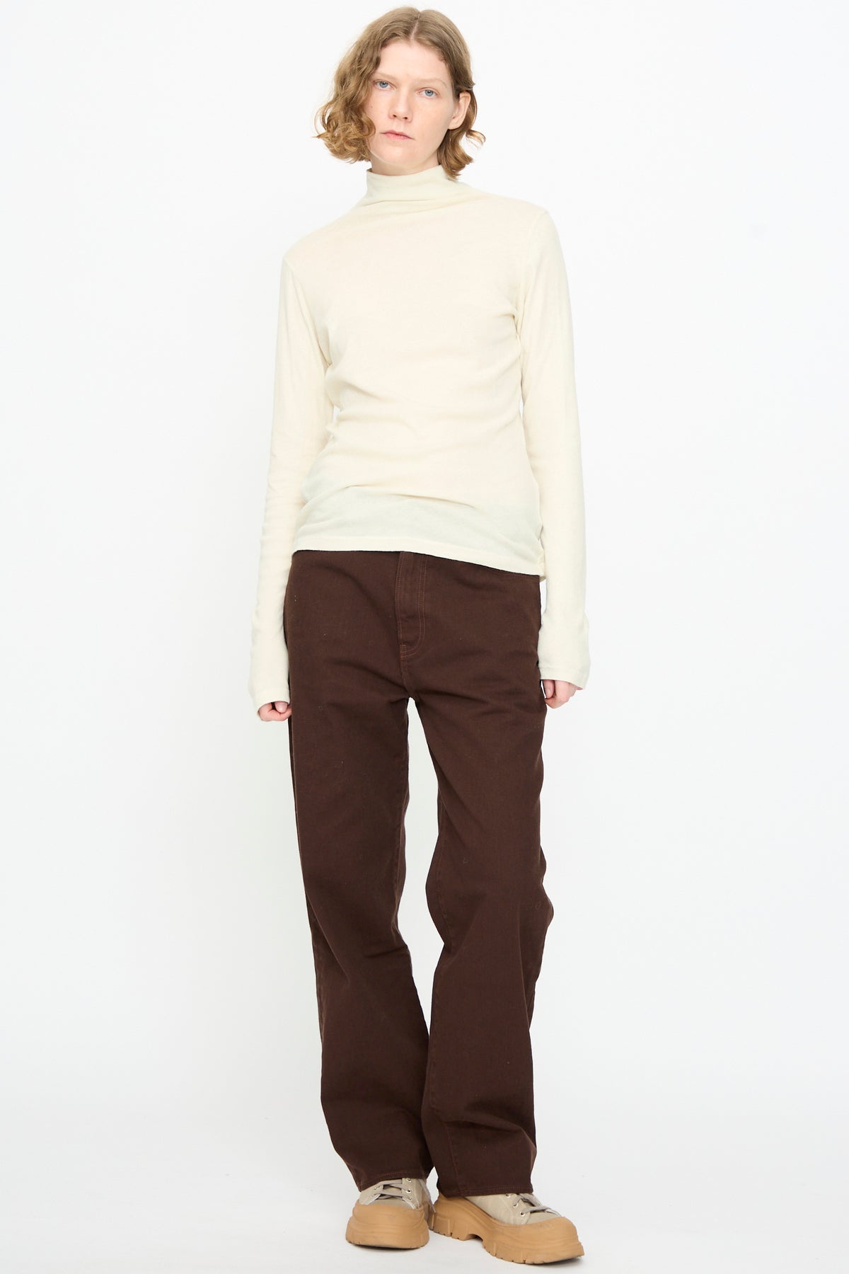 A person stands against a plain white background wearing the Black Crane Turtleneck in Natural, paired with loose brown pants with a relaxed fit and tan shoes.