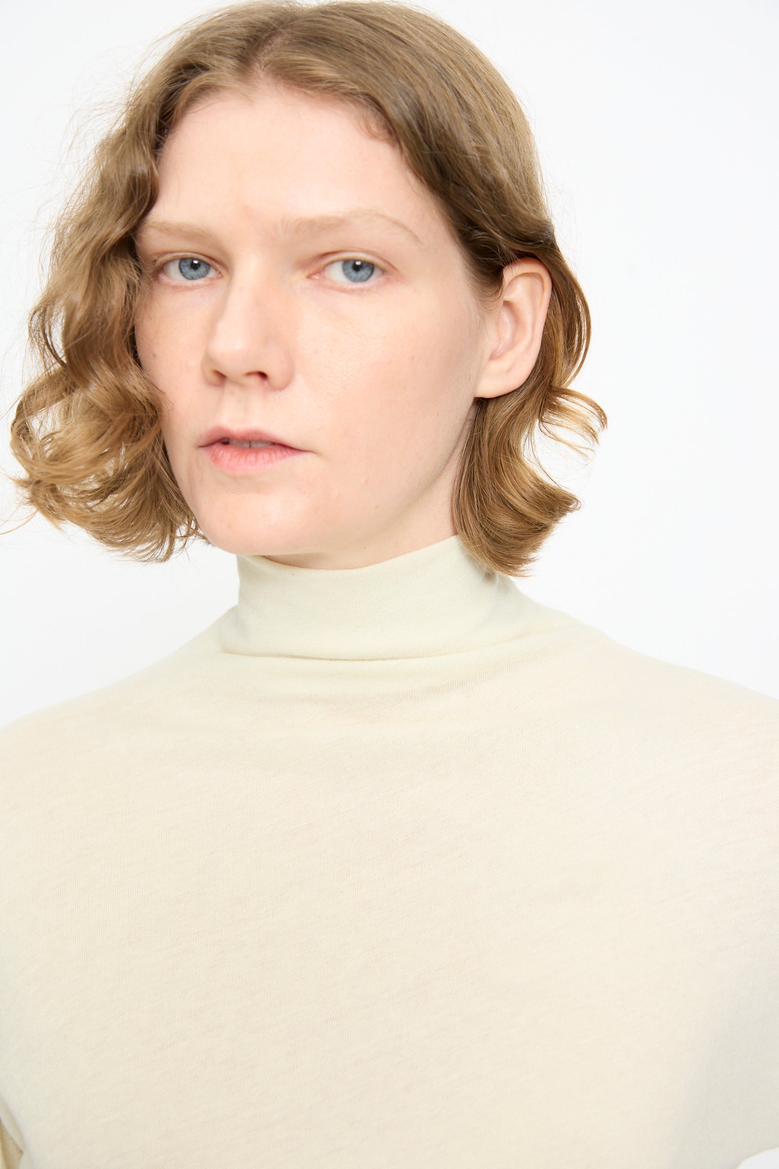 A person with short, wavy light brown hair is wearing the Black Crane Turtleneck in Natural, looking directly at the camera against a plain white background.