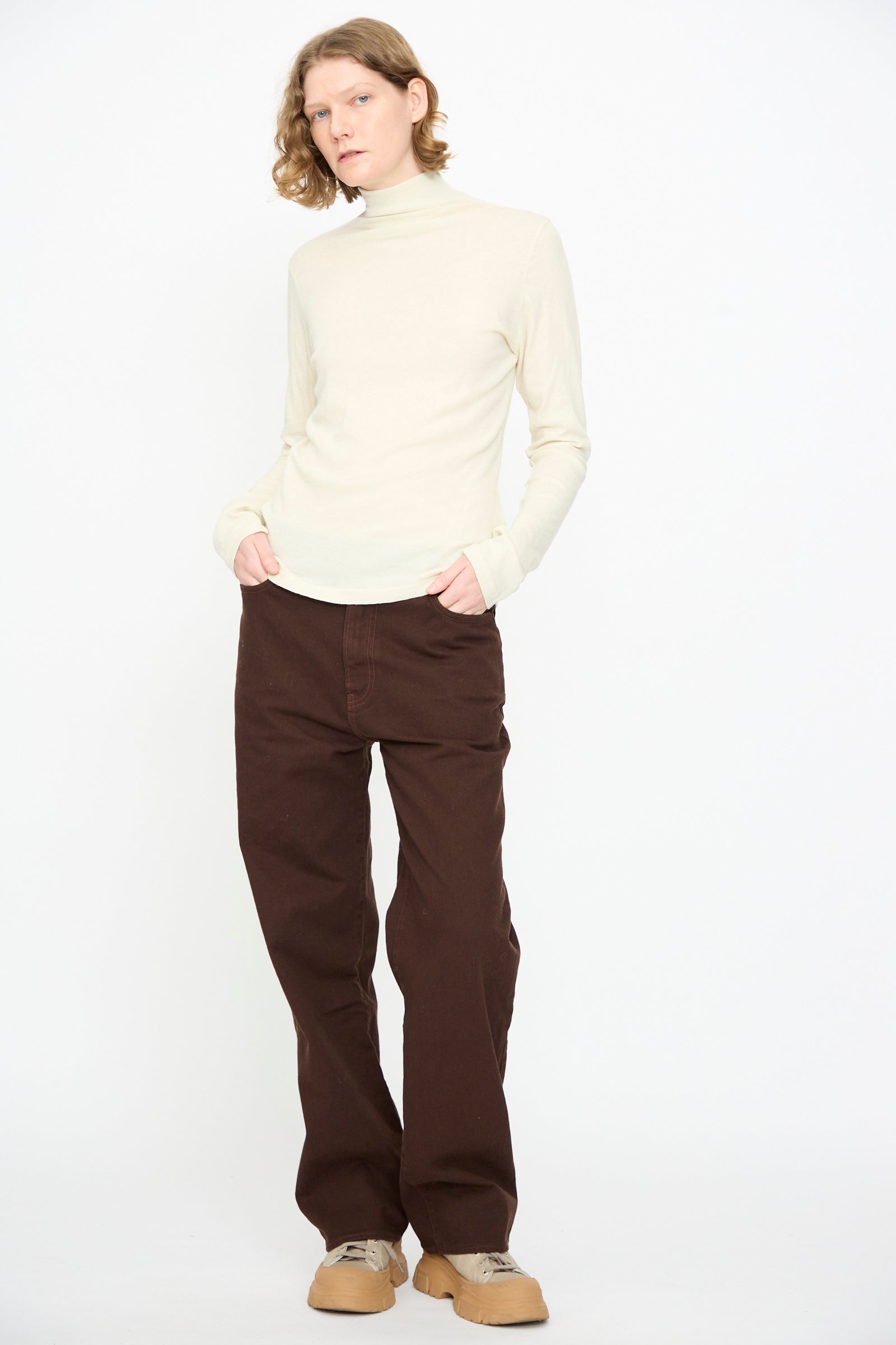 A person stands against a plain white background with hands in pockets, wearing the Black Crane Turtleneck in Natural, brown pants, and beige shoes.