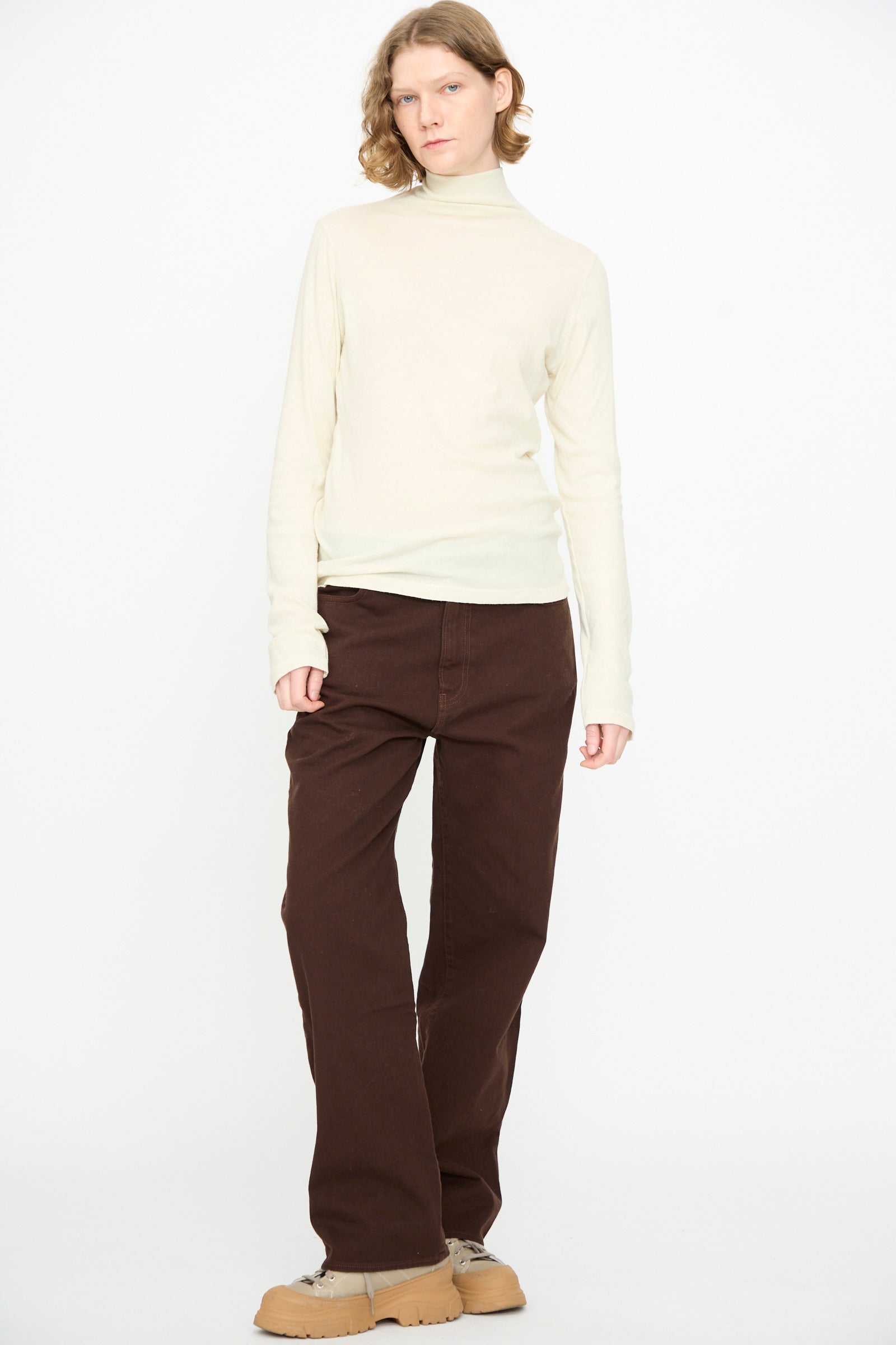 A person stands against a white backdrop, wearing the Black Crane Turtleneck in Natural—a cream cotton-cashmere shirt with a relaxed fit—paired with brown pants and tan shoes.