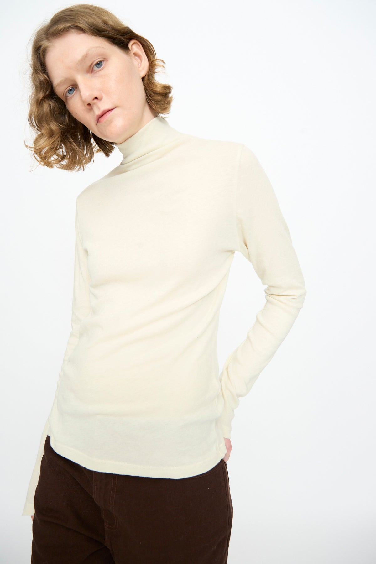 A person with short light brown hair wears the Black Crane Turtleneck in Natural and dark brown pants, standing against a plain white background and looking slightly off-camera in a relaxed fit ensemble.