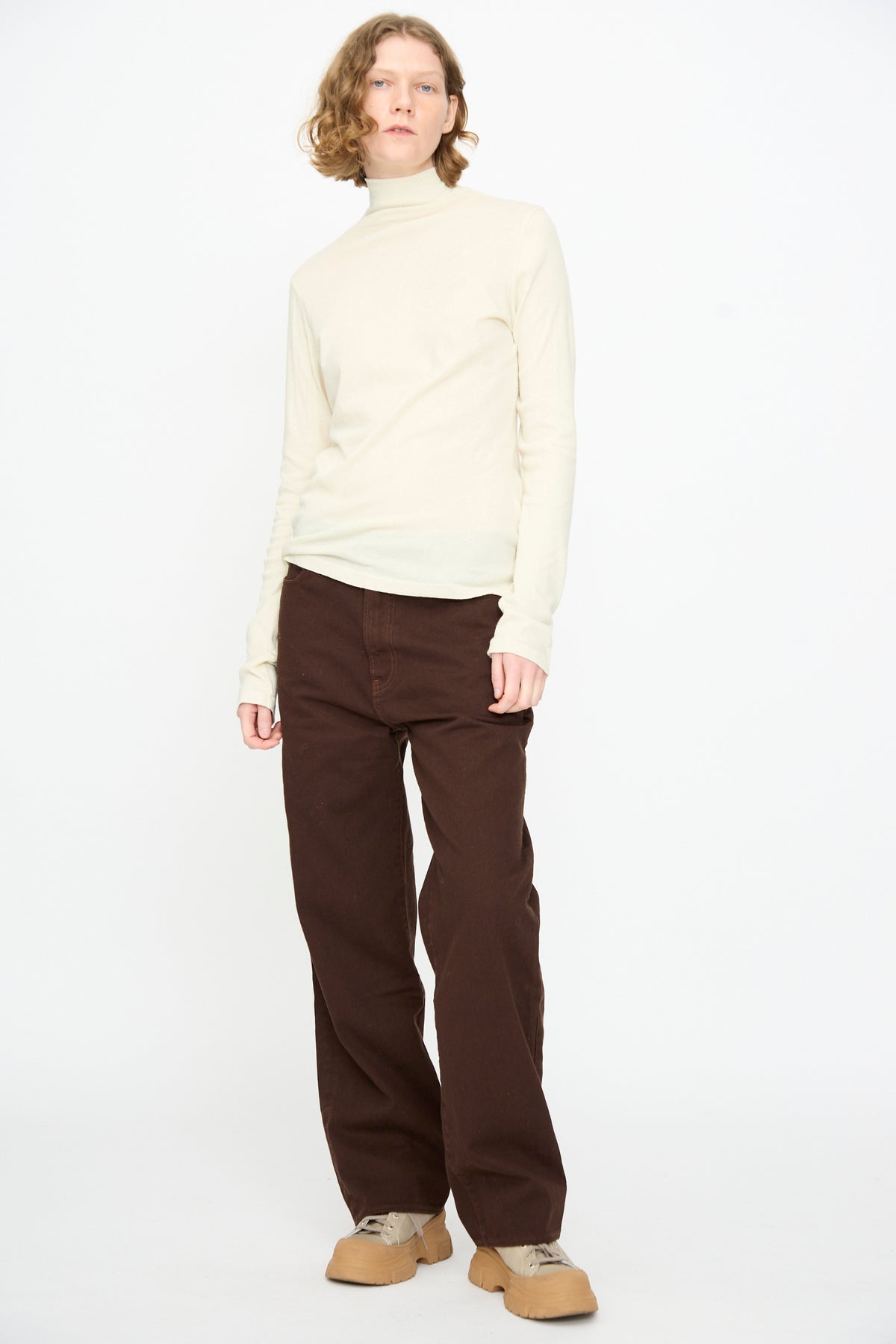 A person stands against a plain background, wearing the Black Crane Turtleneck in Natural, paired with brown pants and tan shoes in a relaxed fit, looking directly at the camera.