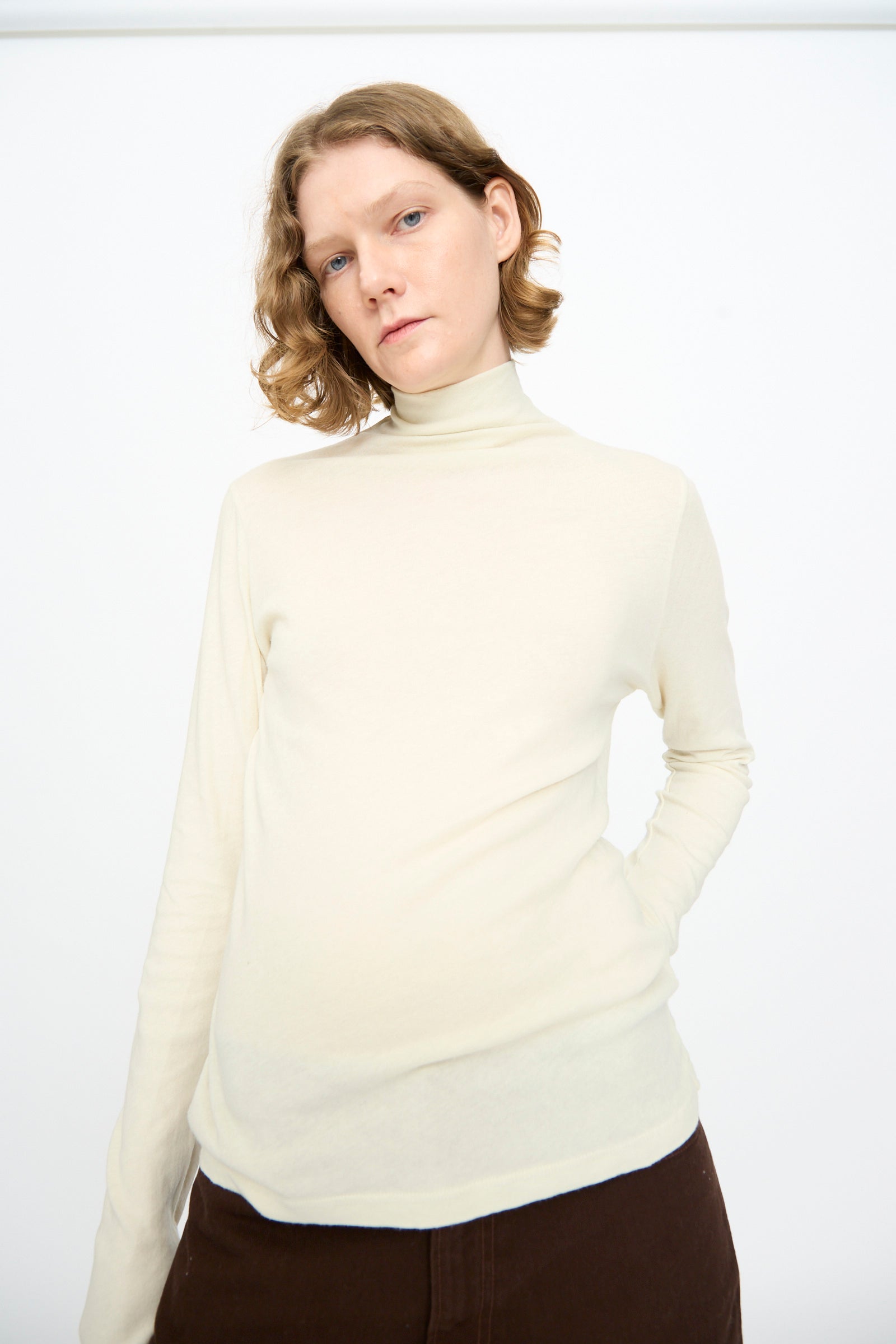 A person with short, wavy hair wears the Black Crane Turtleneck in Natural and relaxed-fit brown pants, standing against a plain white background.