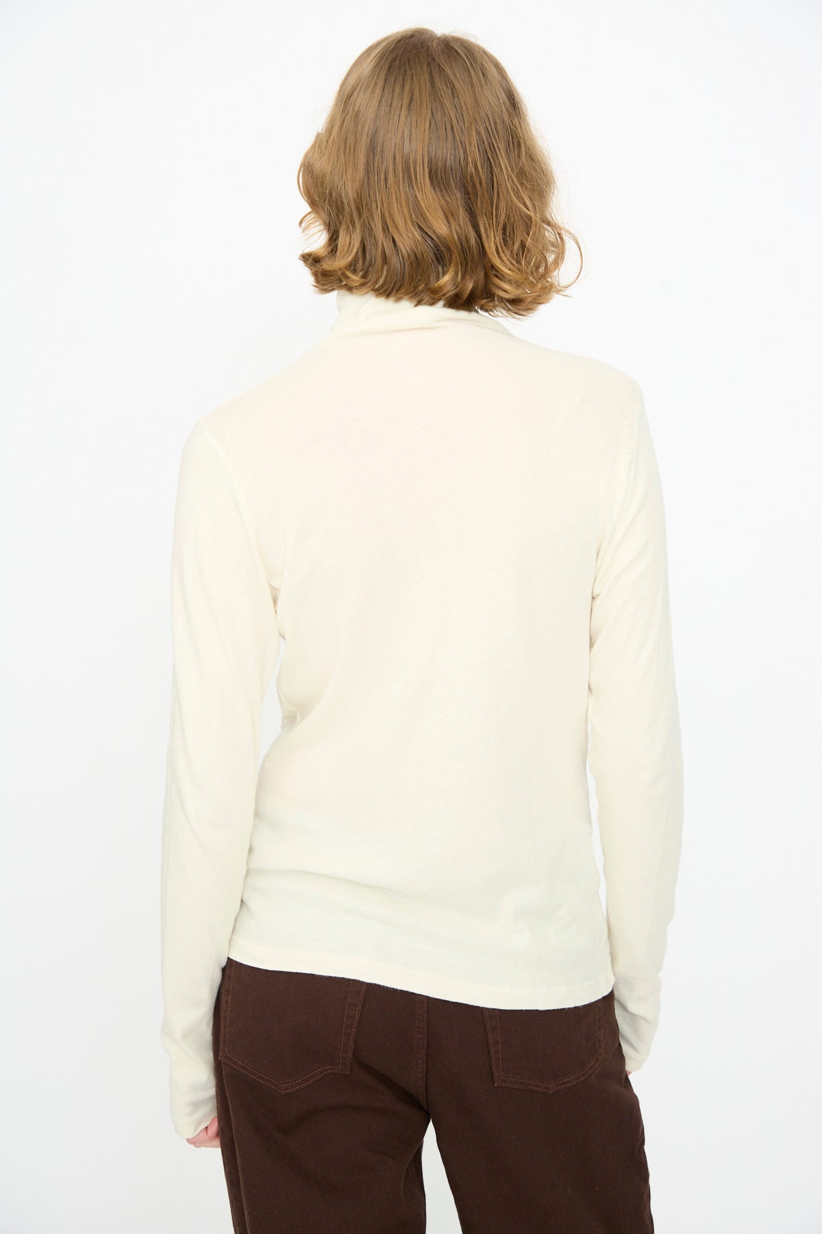 A person with short light brown hair is standing with their back to the camera against a plain white background, wearing the Black Crane Turtleneck in Natural and brown pants.