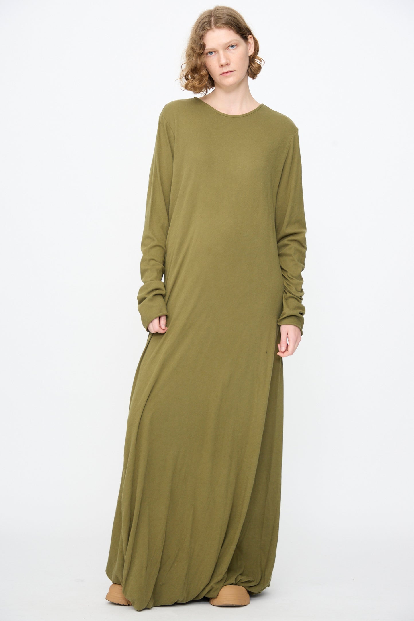 A person stands against a white background wearing the Black Crane Twisted Dress in Army—a loose-fitting, long-sleeve maxi dress crafted from lightweight cotton with an asymmetrical twist drape—paired with tan shoes.