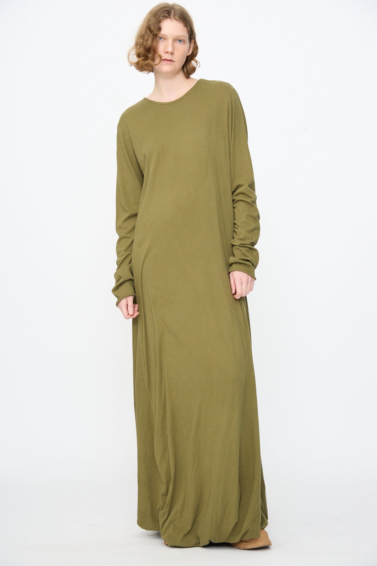 A person stands against a plain background, wearing the Black Crane Twisted Dress in Army—a long sleeve, relaxed-fit maxi dress featuring an asymmetrical twist drape and crafted from lightweight cotton for effortless style.
