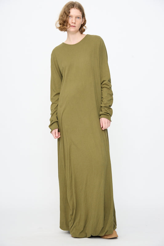 A person stands against a plain background, wearing the Black Crane Twisted Dress in Army—a long sleeve, relaxed-fit maxi dress featuring an asymmetrical twist drape and crafted from lightweight cotton for effortless style.