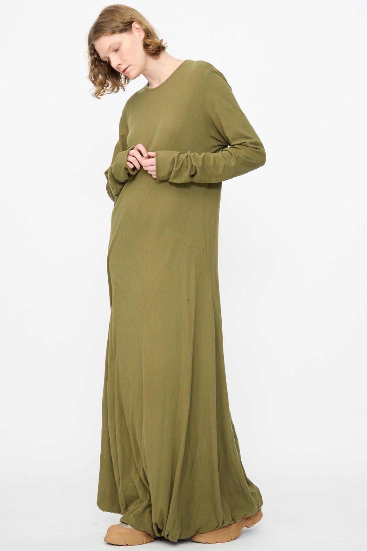 A person stands against a white background wearing the Black Crane Twisted Dress in Army, a long sleeve, loose-fitting maxi dress crafted from lightweight cotton and featuring an asymmetrical twist drape. The dress is paired with tan shoes, while the person’s head is slightly tilted and hands are held close to the torso.