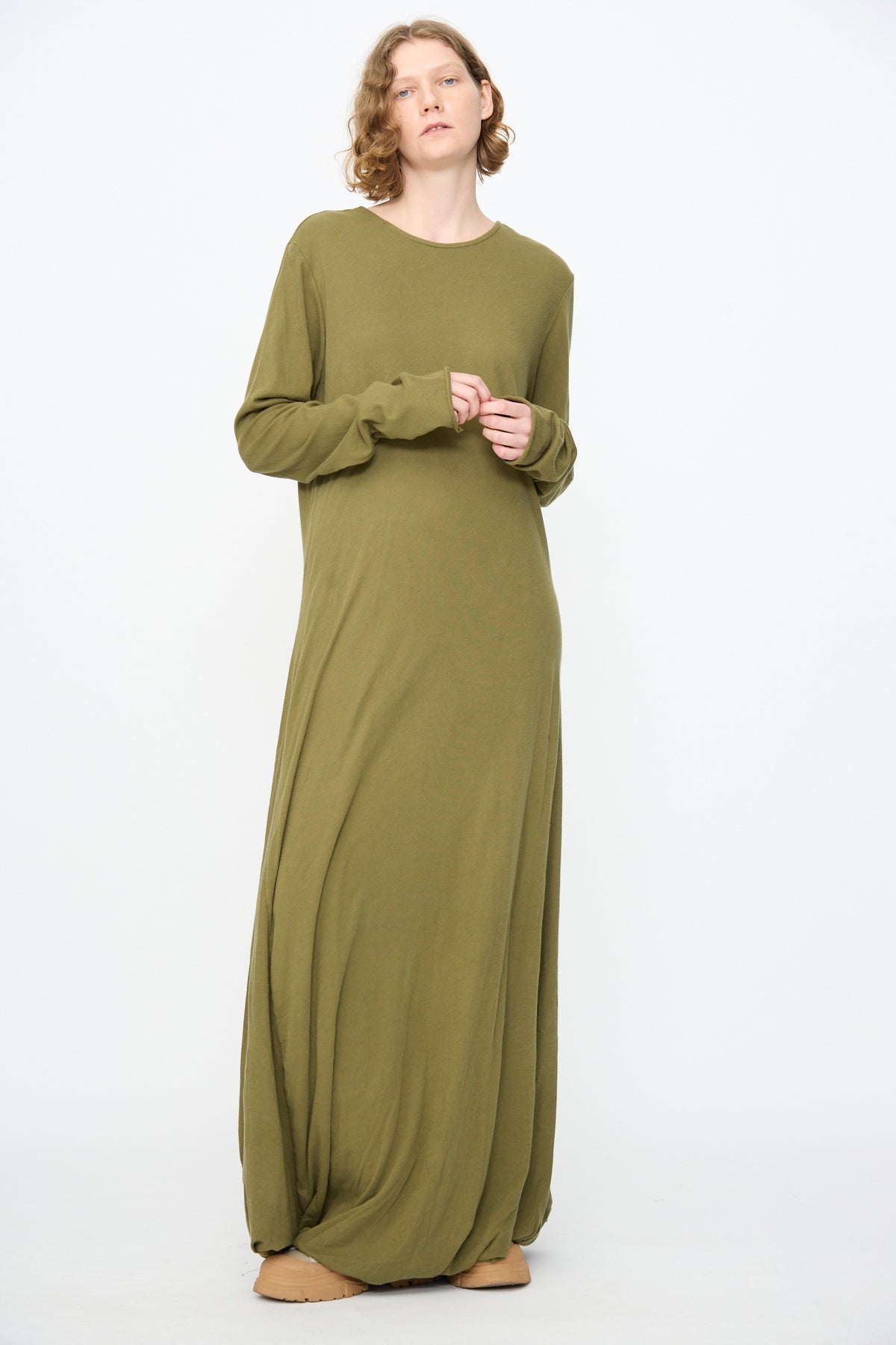 A person stands against a white background, wearing the Black Crane Twisted Dress in Army—a long sleeve maxi dress crafted from lightweight cotton in olive green, featuring an elegant asymmetrical twist drape detail—paired with tan shoes.