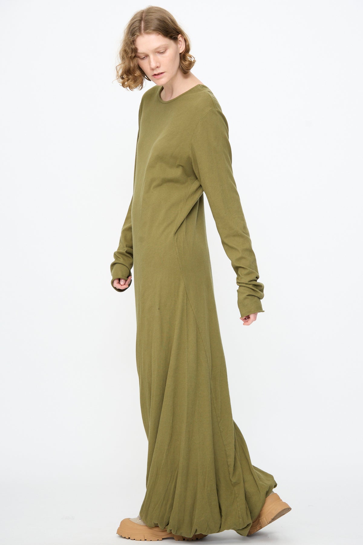 A person wearing the Black Crane Twisted Dress in Army—a loose-fitting, long sleeve maxi dress crafted from lightweight cotton—paired with tan shoes, stands against a plain white background.