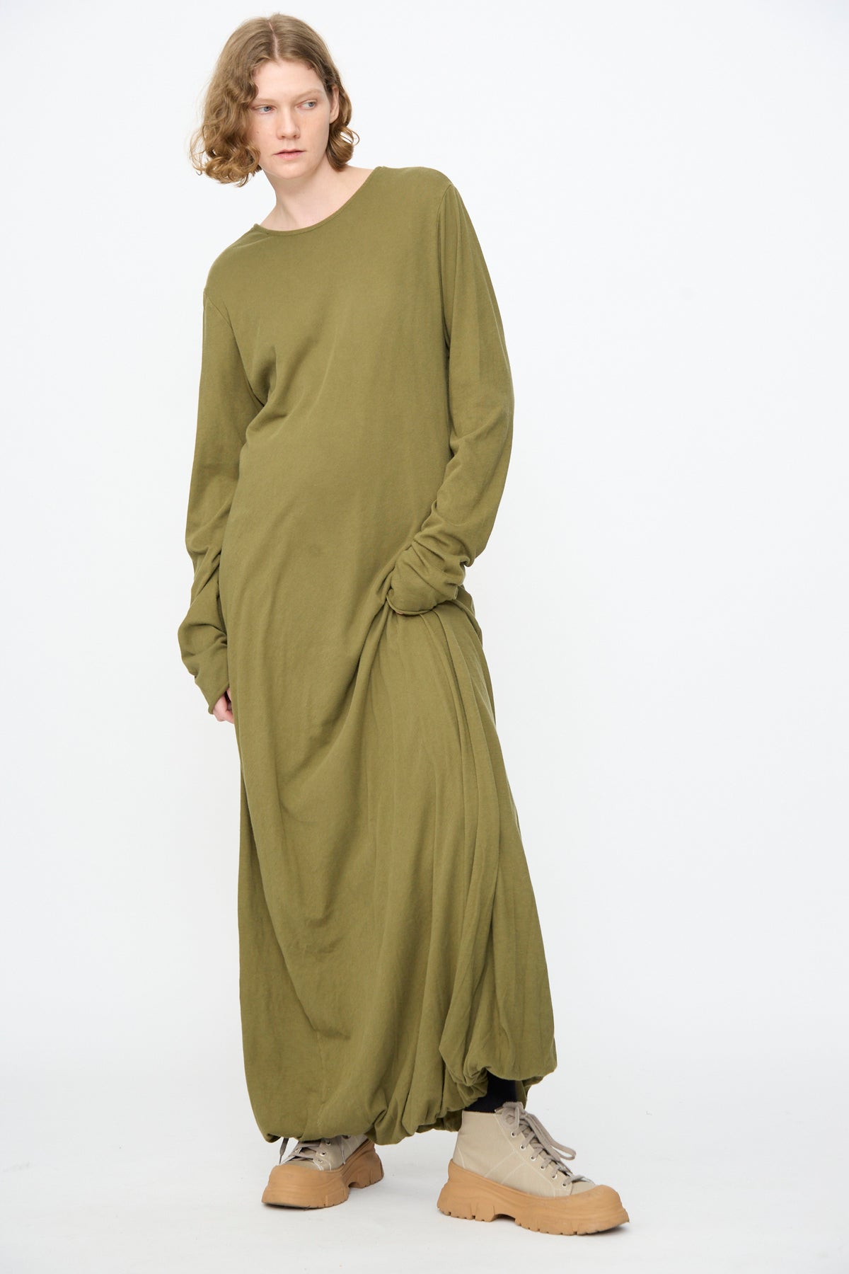 A person with shoulder-length curly hair wears the Black Crane Twisted Dress in Army—a loose, long sleeve maxi dress in lightweight cotton with an asymmetrical twist drape—paired with beige boots and standing against a plain white background.