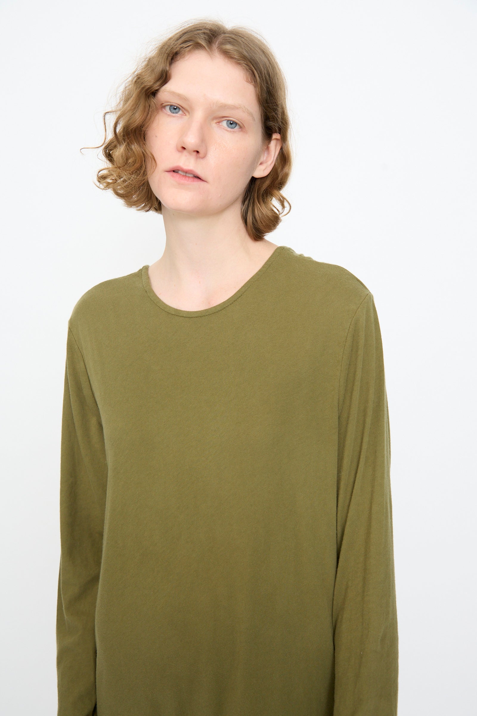 A person with short, wavy light brown hair stands against a plain white background wearing the Black Crane Twisted Dress in Army, an olive green long sleeve maxi dress featuring an asymmetrical twist drape.