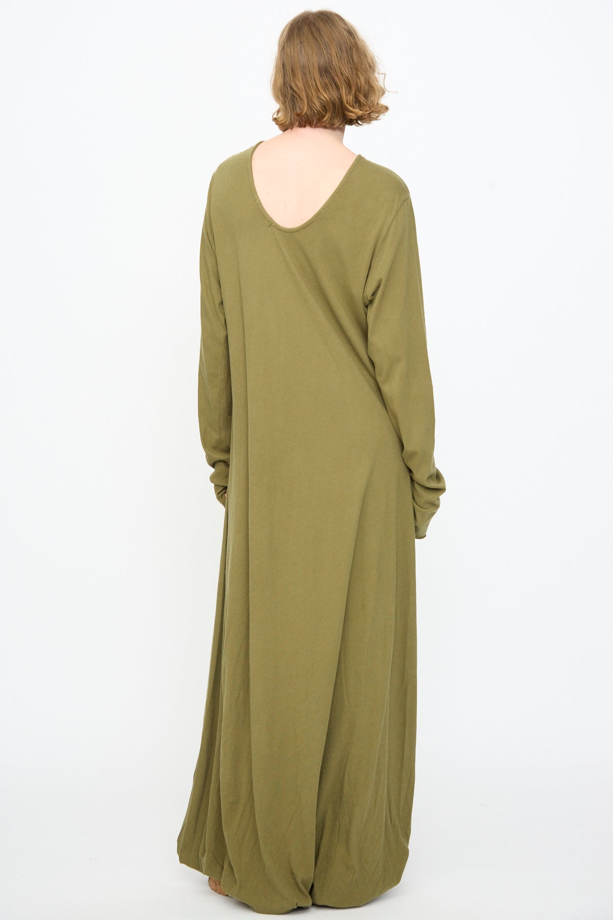 A person with short hair stands barefoot, back facing the camera, wearing the Black Crane Twisted Dress in Army—a floor-length, long sleeve maxi dress in lightweight cotton with an asymmetrical twist drape—against a plain white background.