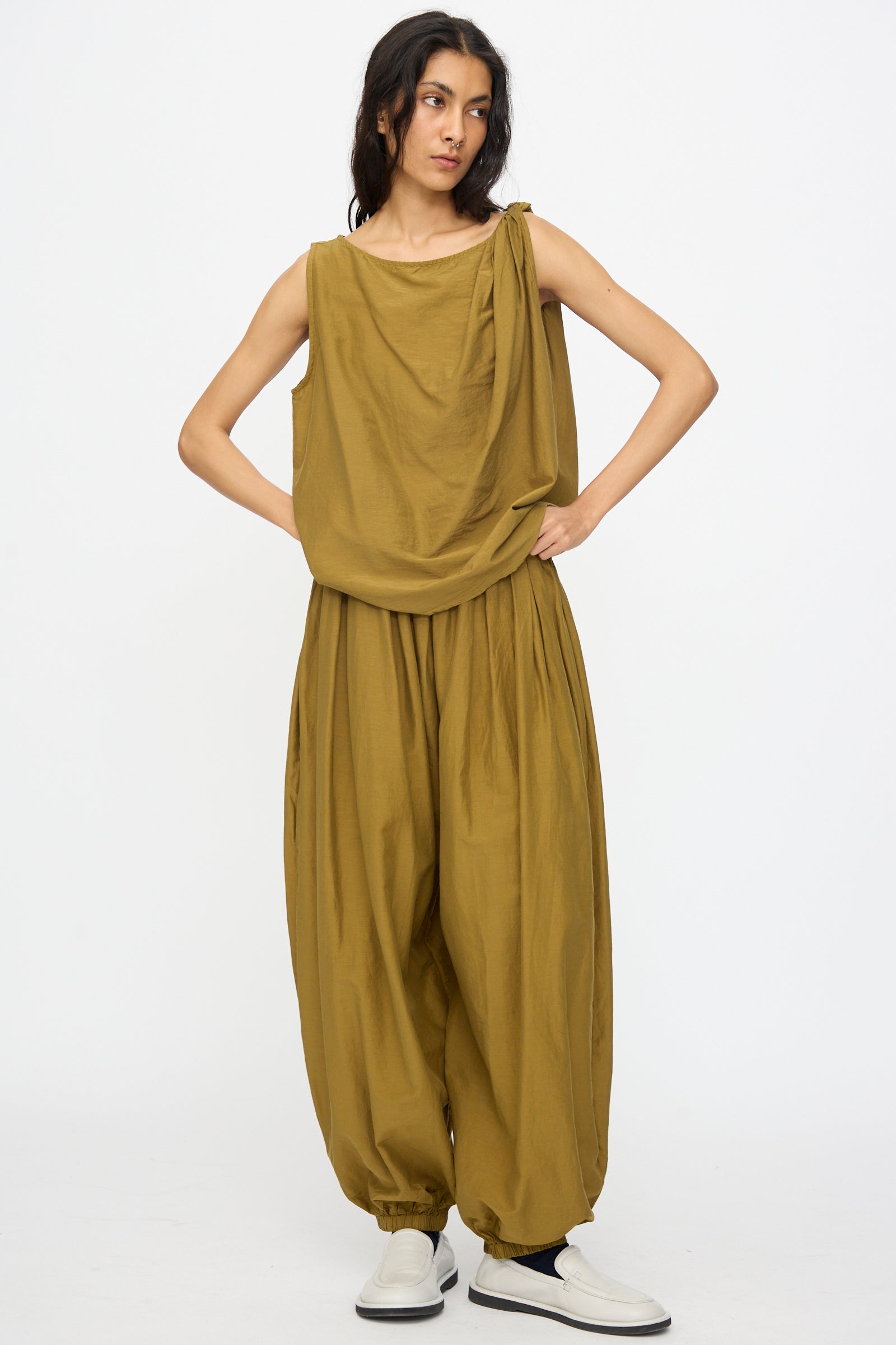 Someone stands against a plain background in the Black Crane Twisted Tank Top in Dark Olive, featuring an asymmetrical twist strap, paired with matching wide, baggy pants and white slip-on shoes.
