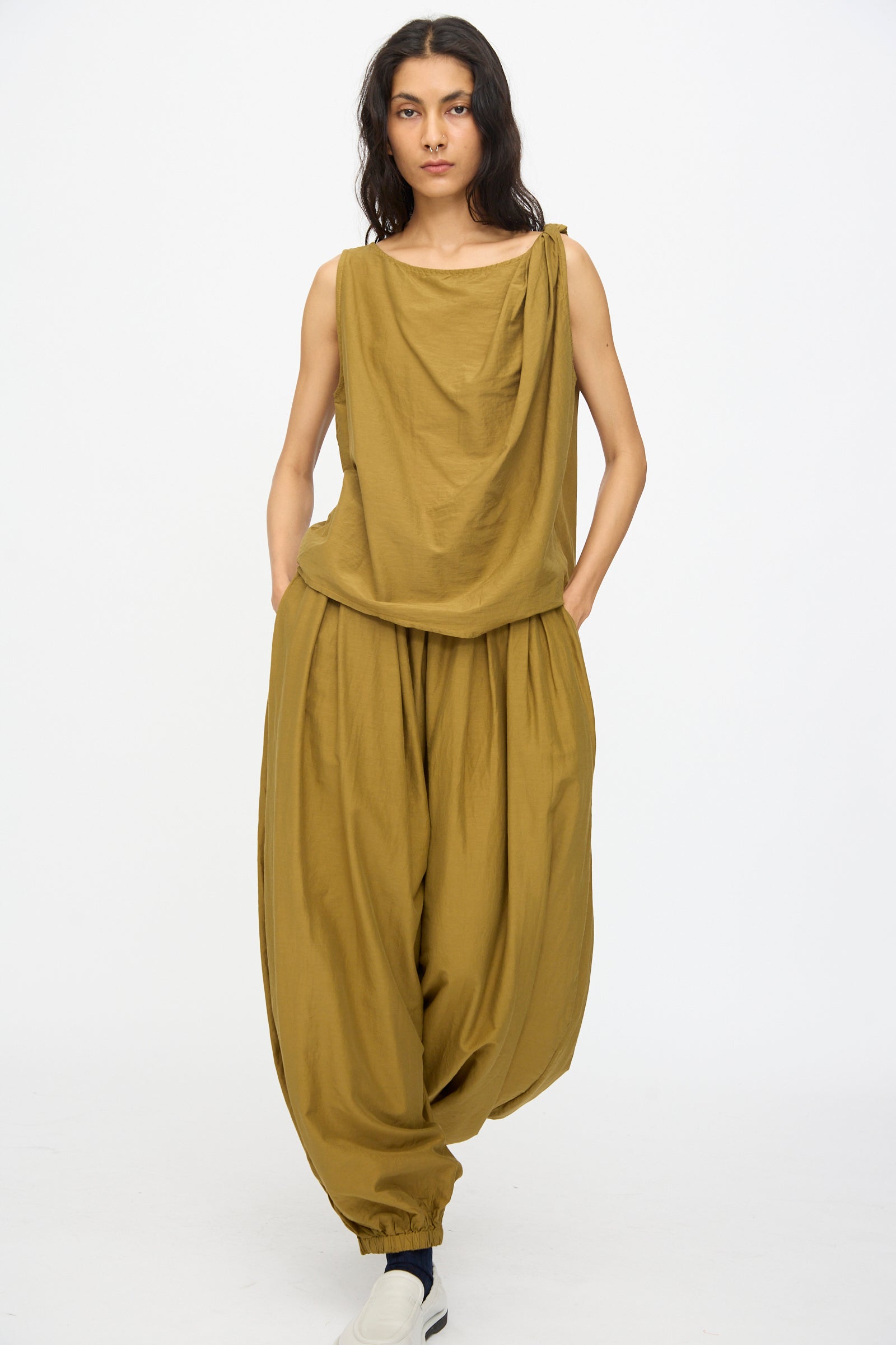 A person wears the Black Crane Twisted Tank Top in Dark Olive, featuring an asymmetrical twist strap, paired with matching wide gathered pants, against a plain white background.
