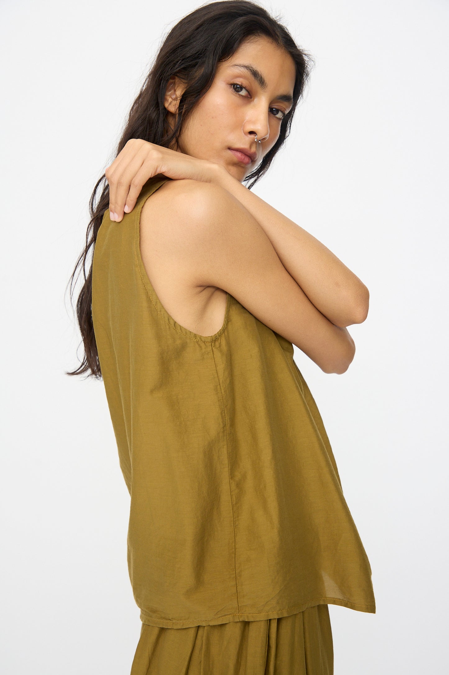 A person with long dark hair poses against a plain background, wearing the Black Crane Twisted Tank Top in Dark Olive with asymmetrical twist straps and matching bottoms, one arm bent and looking at the camera.