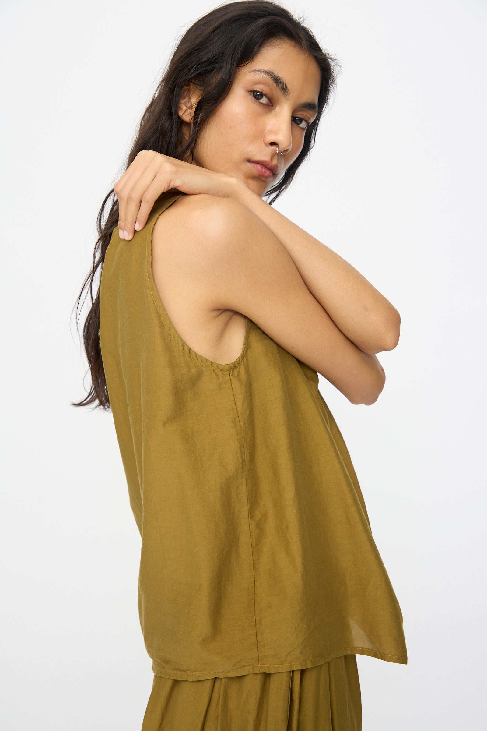 A person with long dark hair poses against a plain background, wearing the Black Crane Twisted Tank Top in Dark Olive with asymmetrical twist straps and matching bottoms, one arm bent and looking at the camera.