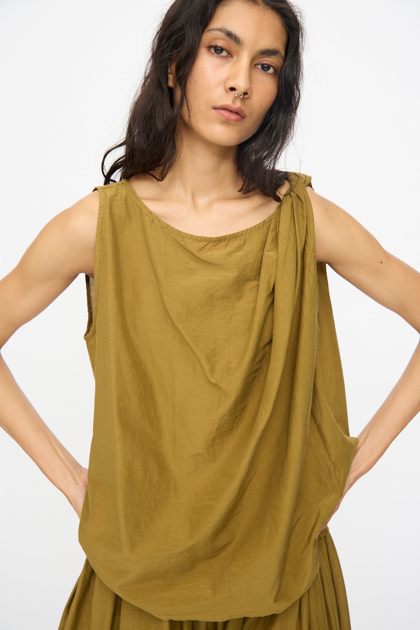 A person with long dark hair wears the Black Crane Twisted Tank Top in Dark Olive and a matching skirt, standing against a plain light background.