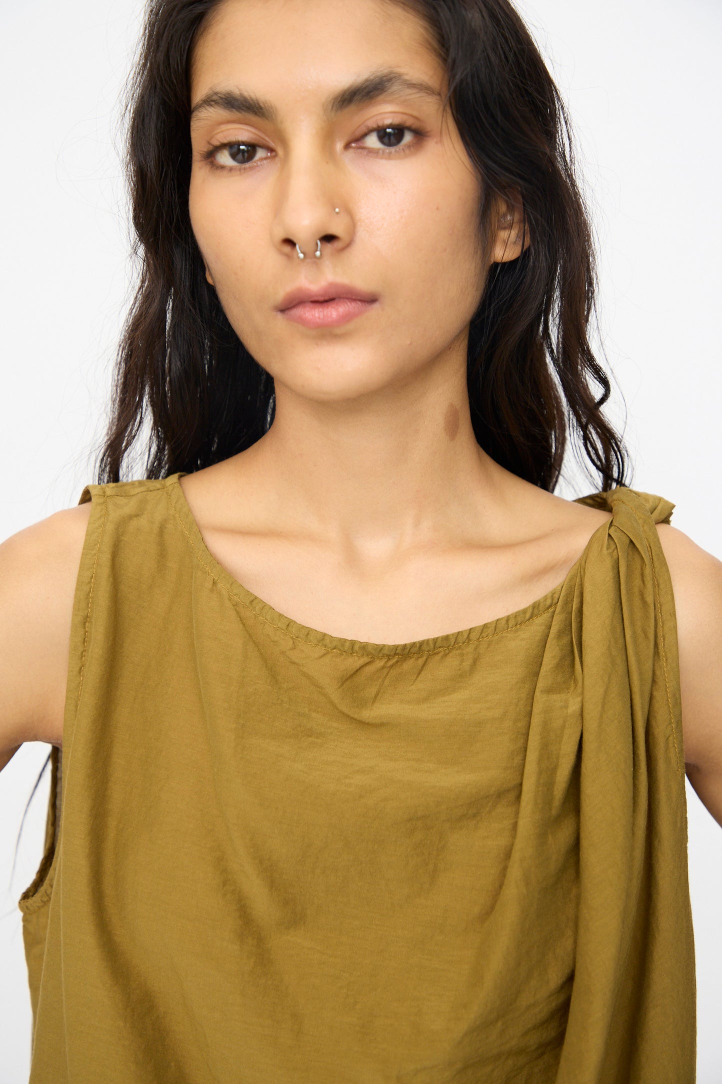 A person with long dark hair models the Black Crane Twisted Tank Top in Dark Olive, featuring an asymmetrical twist strap, against a plain light background.