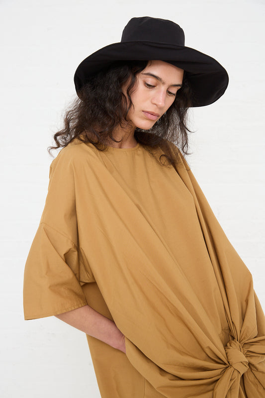 Woman in a stylish oversized Black Crane beige tunic and Black Crane Cotton Wavy Hat in Black standing against a white wall, with her head tilted downward.