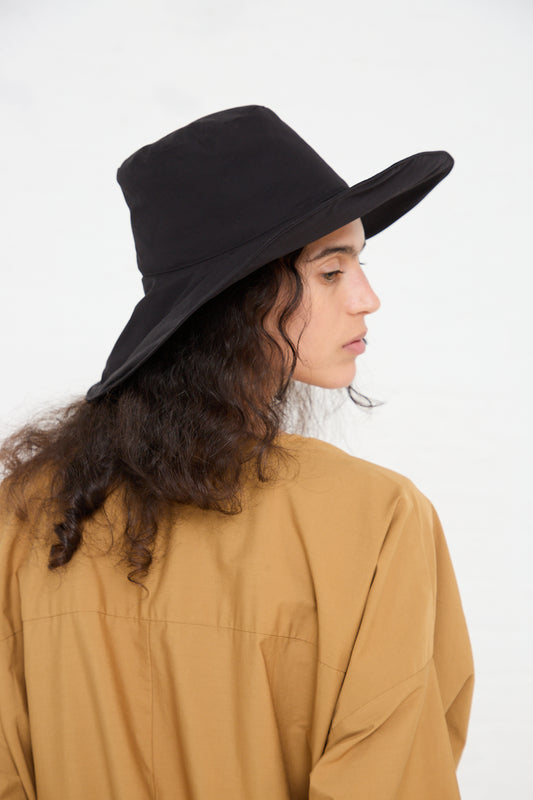 Profile view of a person with curly hair wearing a Black Crane Cotton Wavy Hat in Black and a beige shirt, against a plain white background.