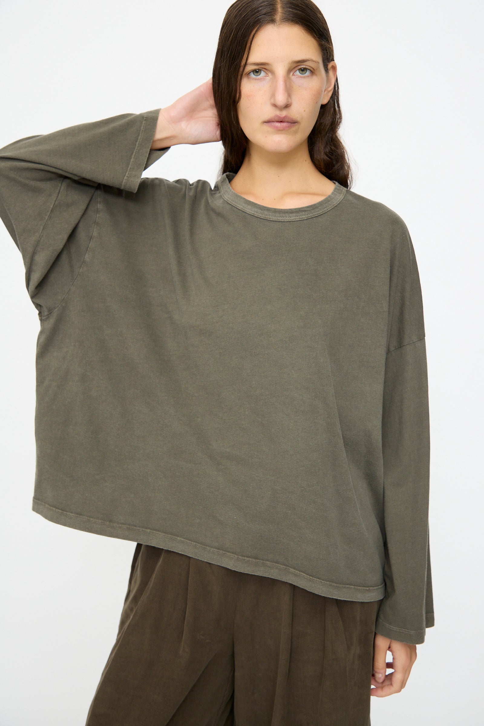 A person with long brown hair wears the Black Crane Wide Long Sleeve Tee in Slate and brown pants, standing against a plain white background.