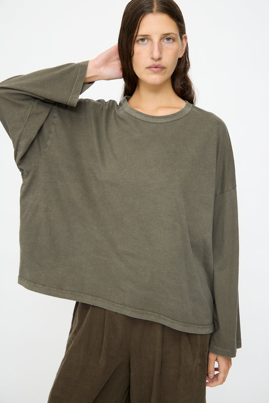 A person with long brown hair wears the Black Crane Wide Long Sleeve Tee in Slate and brown pants, standing against a plain white background.