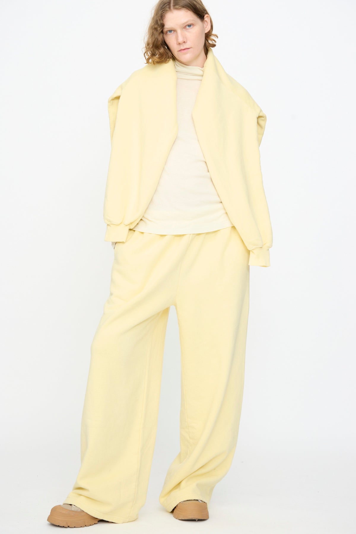 A person stands against a white background wearing Black Crane’s Wide Long Sweatpants in Custard—a loose, pale yellow cotton-cashmere style—paired with a matching jacket draped over their shoulders, a beige top, and tan shoes.