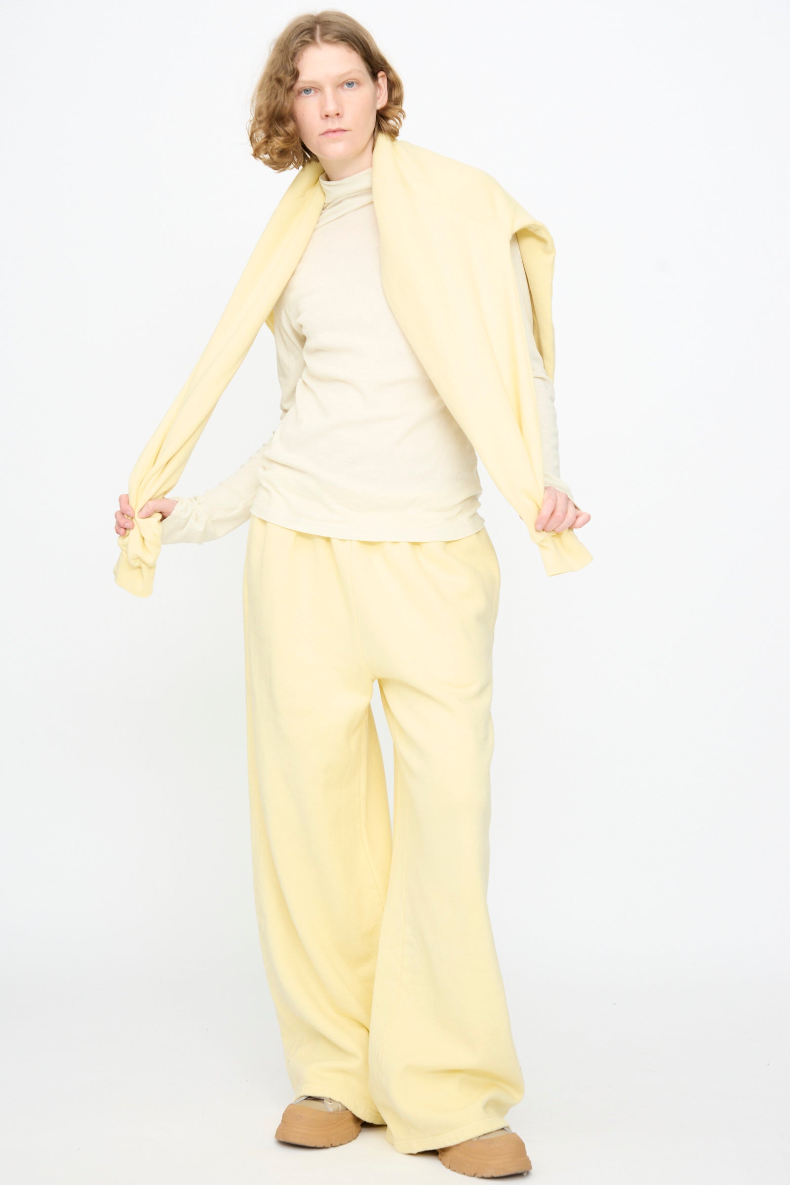 A person stands against a white background, wearing the Black Crane Wide Long Sweatpants in Custard, paired with a long-sleeve shirt, tan shoes, and a sweater draped over their shoulders while holding the sweater’s sleeves.