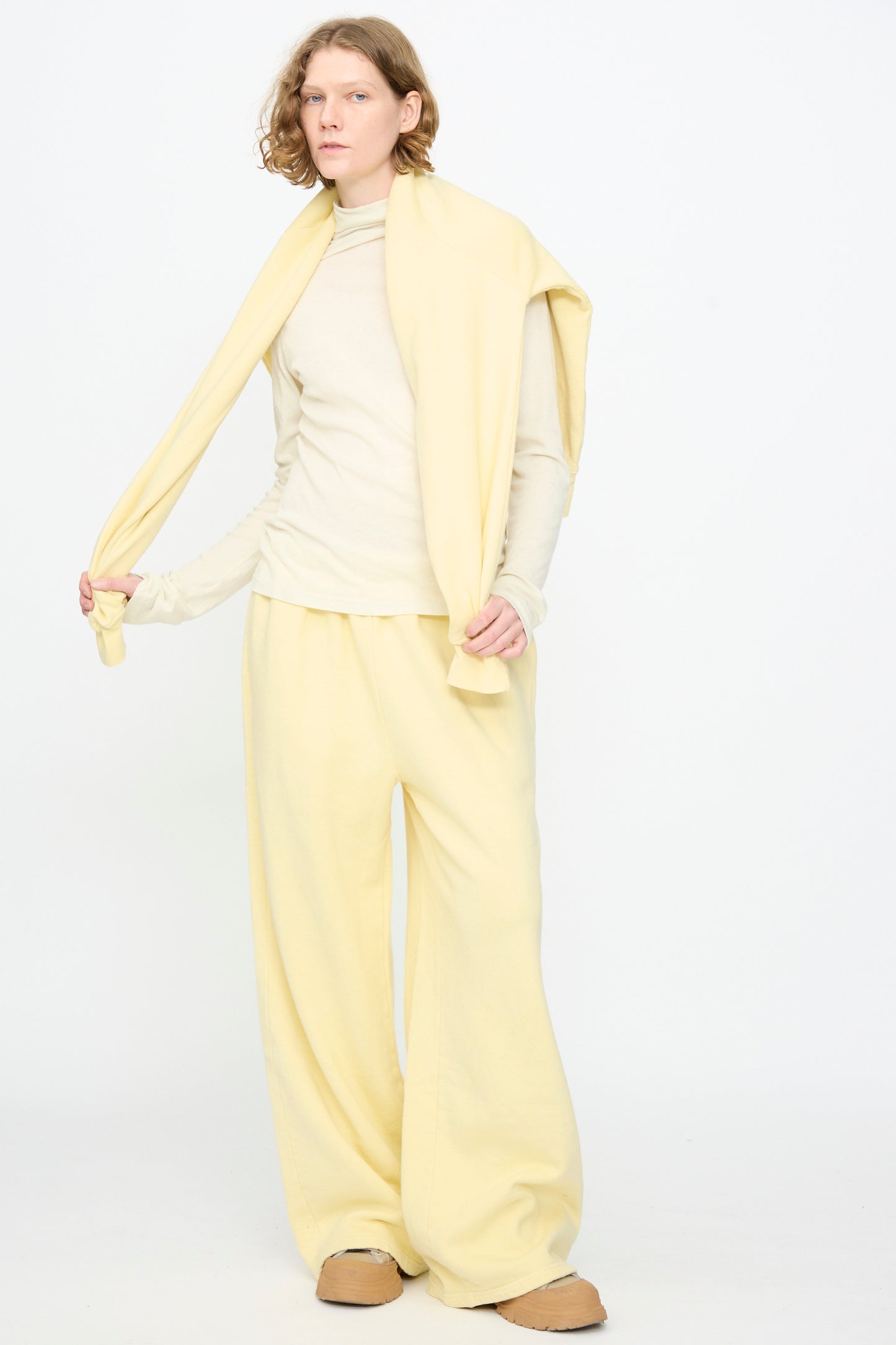 A person stands against a plain white background wearing Black Crane’s Wide Long Sweatpants in Custard—a relaxed, pale yellow style—paired with a cream long-sleeve top, tan slip-on shoes, and a yellow sweater draped over their shoulders.