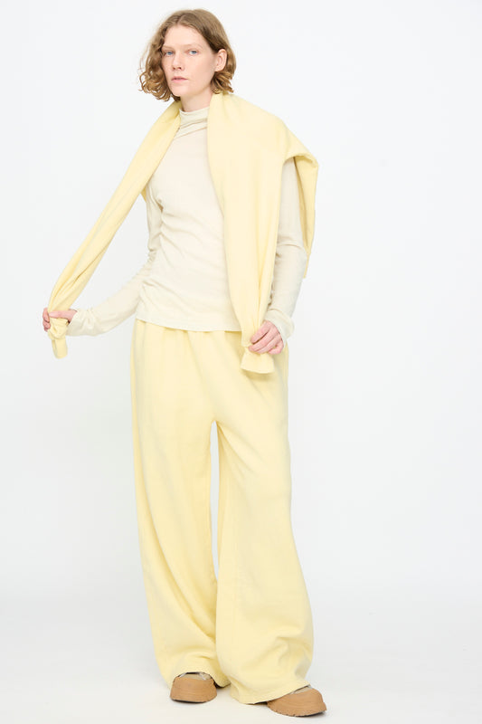 A person stands against a plain white background wearing Black Crane’s Wide Long Sweatpants in Custard—a relaxed, pale yellow style—paired with a cream long-sleeve top, tan slip-on shoes, and a yellow sweater draped over their shoulders.