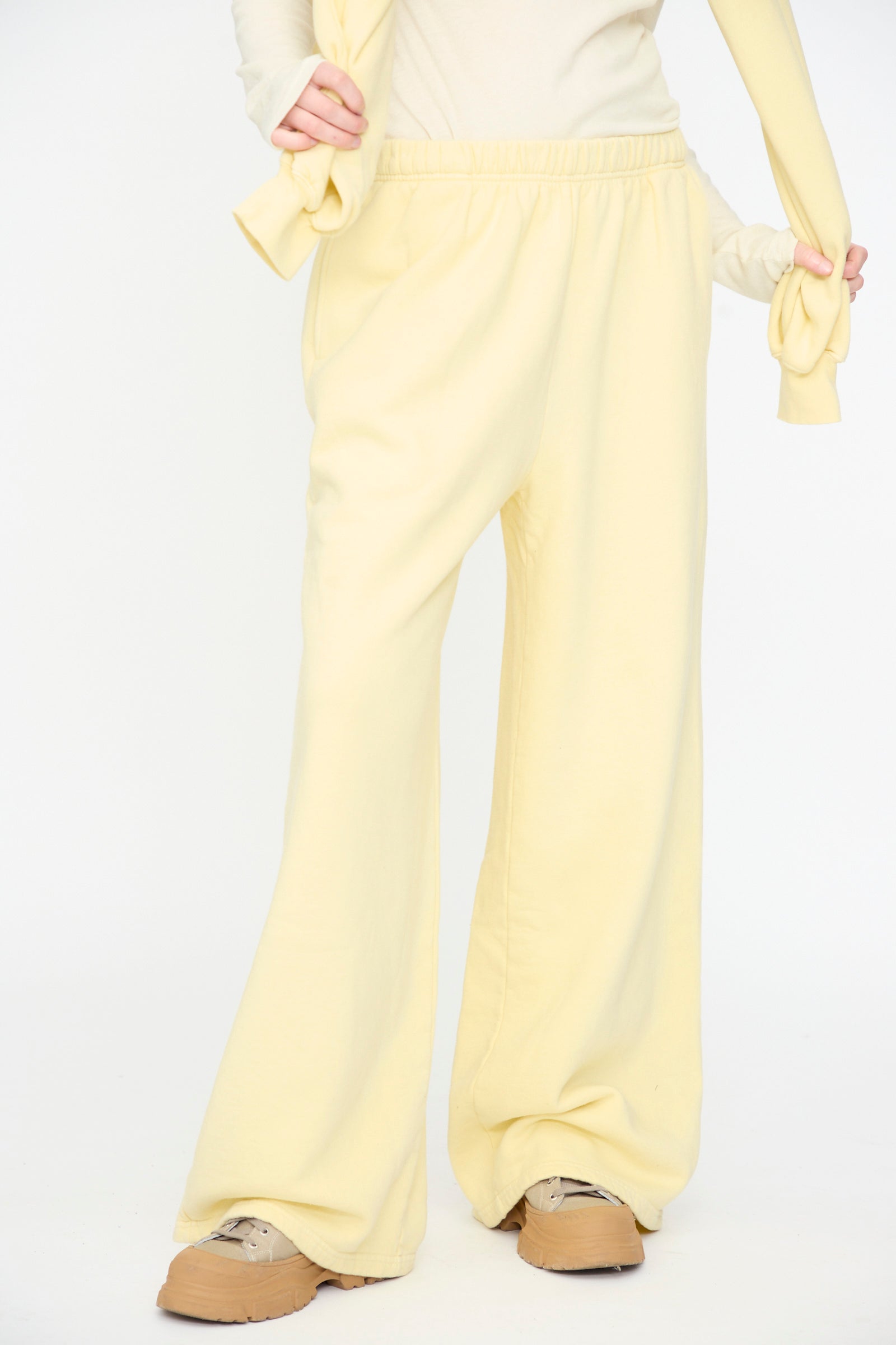 A person is shown from the shoulders down, wearing Black Crane’s Wide Long Sweatpants in Custard paired with tan sneakers, against a plain white background.