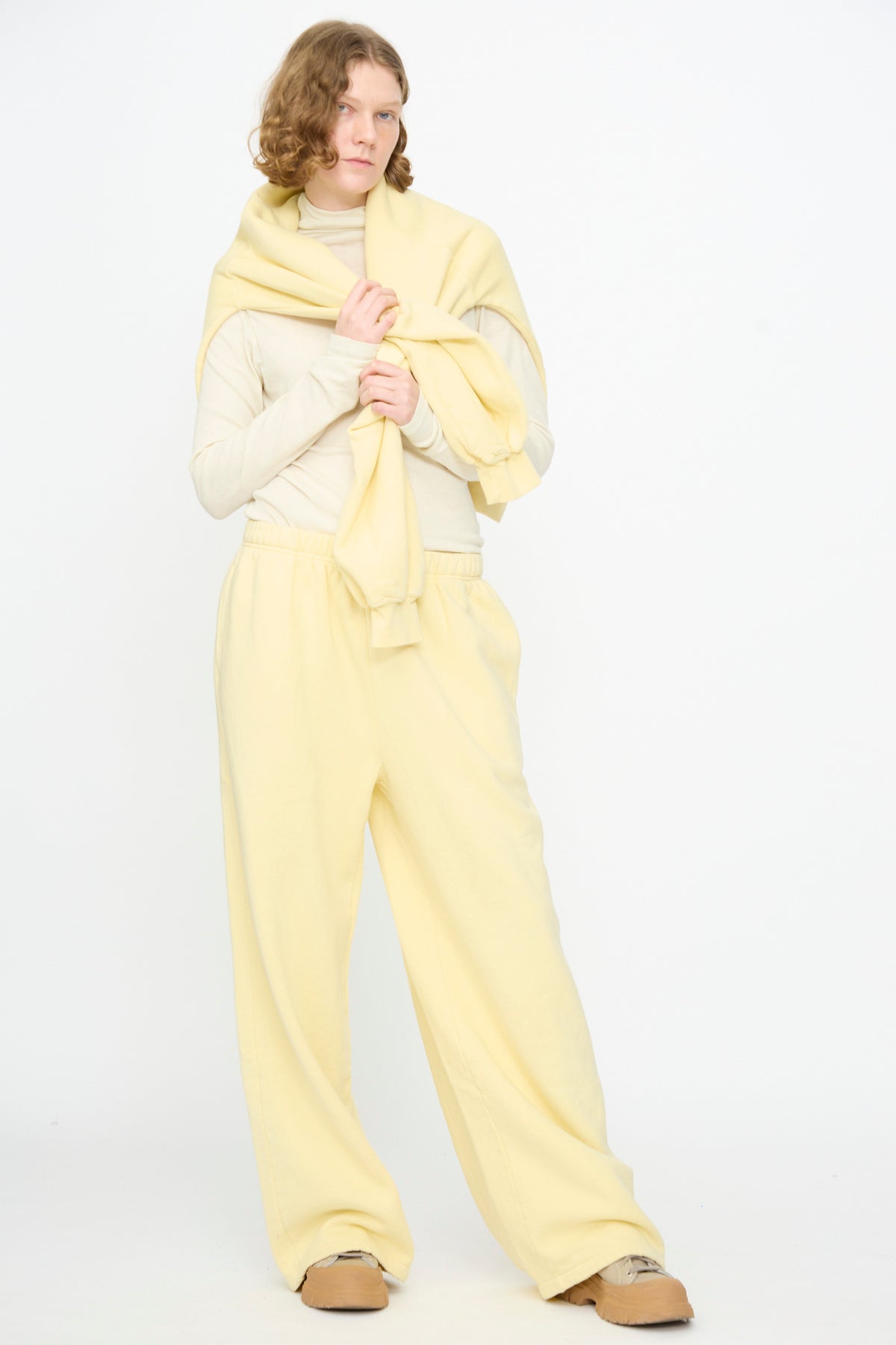 A person with shoulder-length hair wearing a cream turtleneck, a yellow sweatshirt draped over their shoulders, Black Crane Wide Long Sweatpants in Custard, and tan shoes stands against a plain white background.