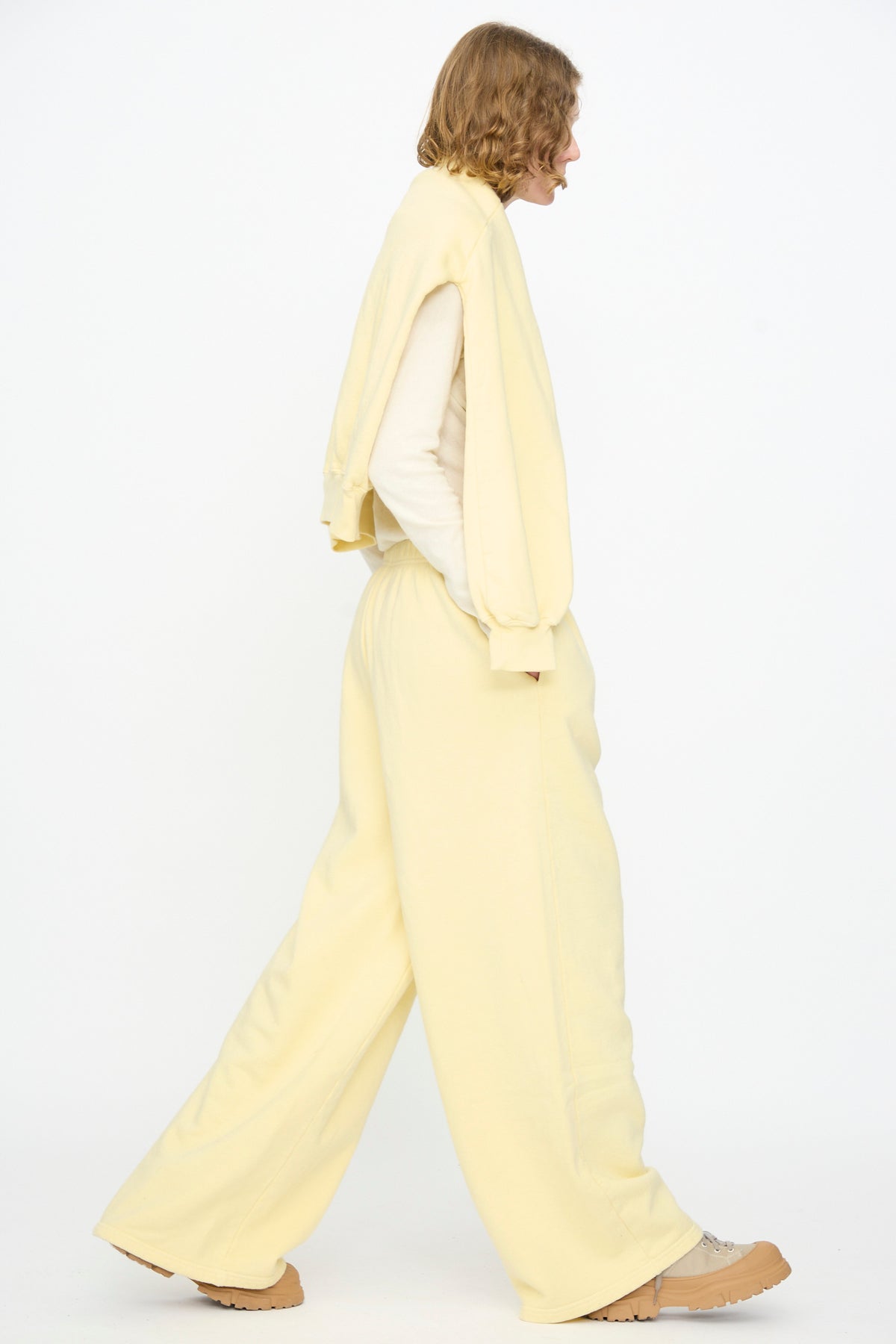 A person in side profile is wearing a cream-colored sweatshirt draped over their shoulders, paired with Black Crane’s Wide Long Sweatpants in Custard, featuring a relaxed fit and wide-leg silhouette, along with beige shoes, walking against a plain white background.